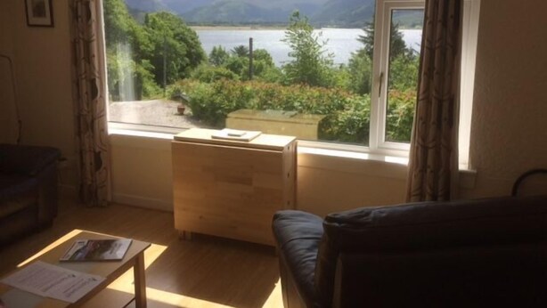 Quarto com uma grande janela que oferece vista para um lago e montanhas. Há uma mesa de madeira e um sofá preto.