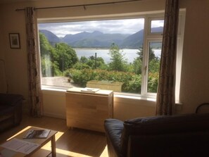 A room with a large window offering a view of a lake and mountains. There is a wooden table and a black sofa. - Old Springwell Croft (Fort William)