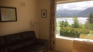 Sala de estar com sofá de couro preto, mesinha de madeira e janela com vista para um lago e montanhas.
