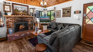 Ein gemütliches Blockhaus-Interieur mit Kamin, Ledersofa, Holzbalkendecke und verschiedenen gerahmten Bildern.