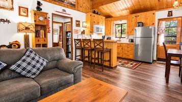 A cozy cabin interior with a wooden ceiling, a kitchenette, a dining area, and a living room with a sofa and a coffee table.