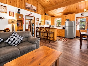 A cozy cabin interior with a wooden ceiling, a kitchenette, a dining area, and a living room with a sofa and a coffee table. - The Cuddle Inn (Big Bear Lake)