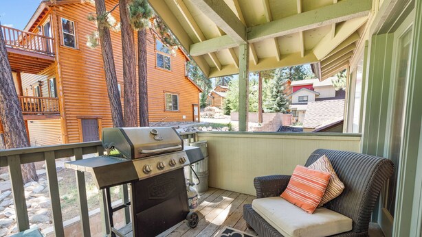 A covered outdoor area with a grill, a wicker sofa with cushions, and a view of a wooden house. - Bear Mountain Escape Condo (Big Bear Lake)