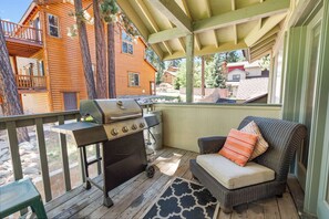 Un espace extérieur abrité, doté d’un barbecue, d’un canapé en osier avec des coussins et offrant une vue sur une maison en bois.