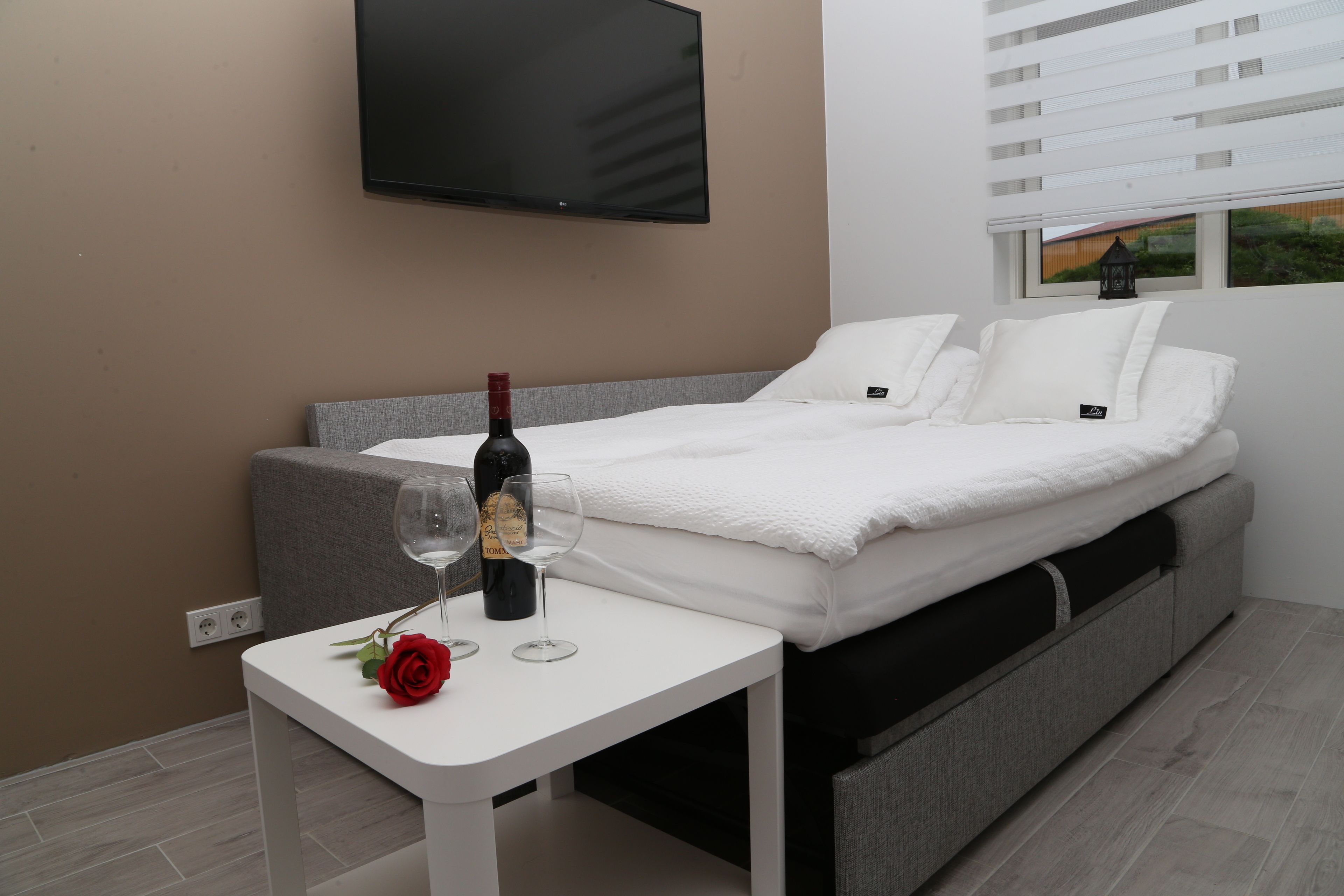 Un dormitorio moderno con una cama, una mesita con una botella de vino y una copa, y un televisor montado en la pared.