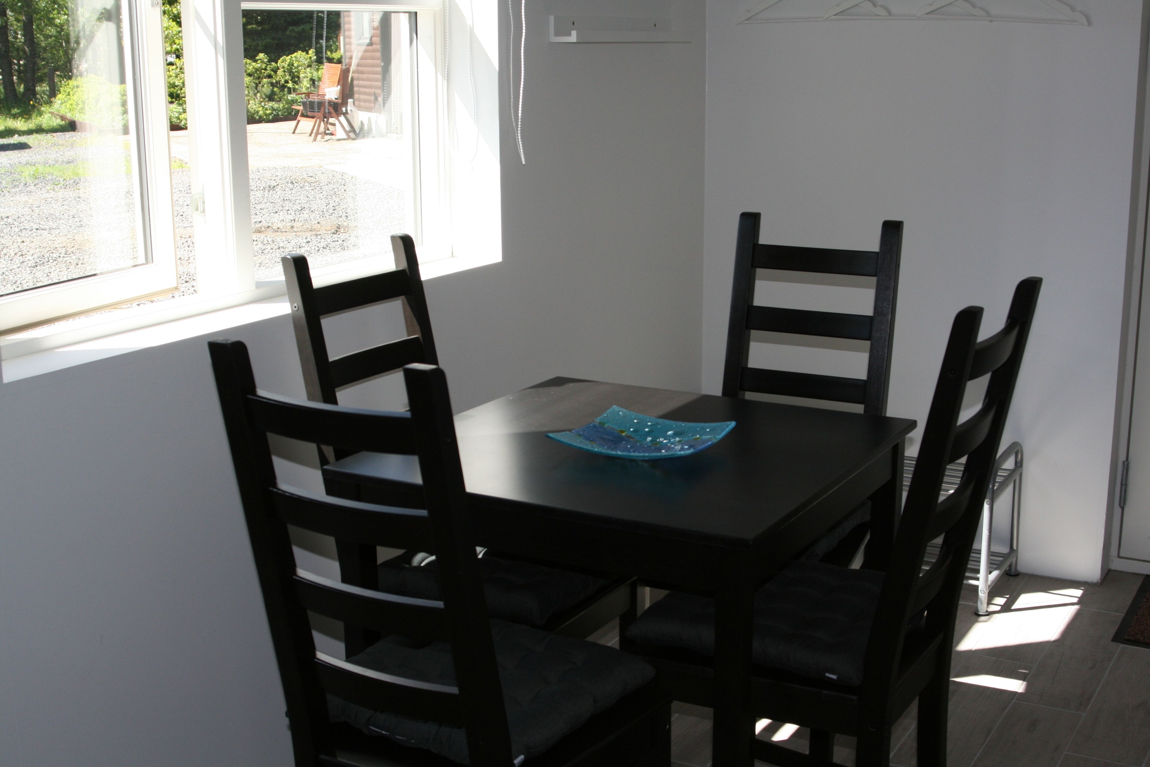 A dining area with a table and four chairs, a blue decorative item on the table, and a view of the outdoors through a window.
