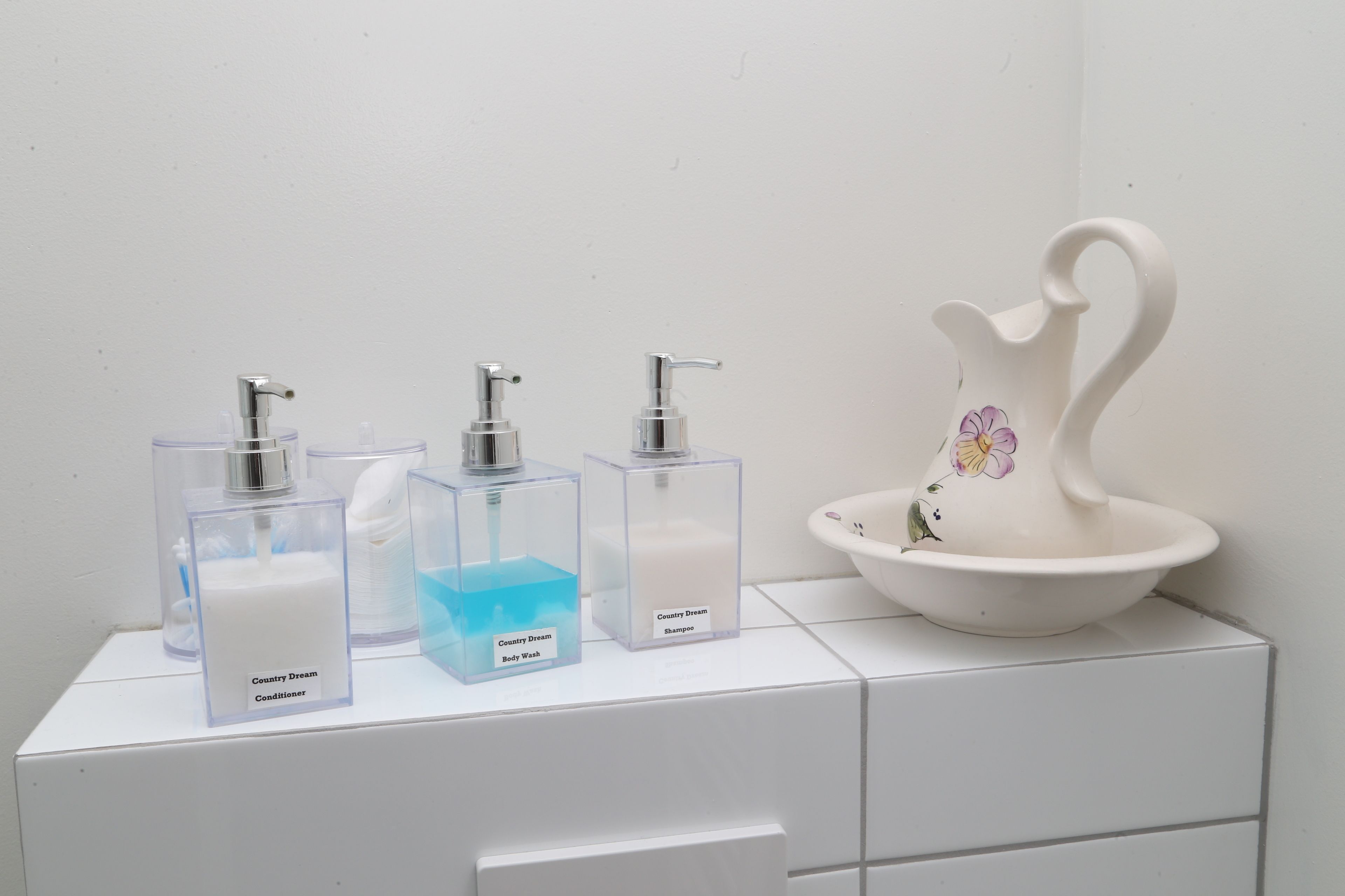 A bathroom with a white sink, three soap dispensers labeled 'Country Dream Body Wash,' 'Country Dream Shampoo,' and 'Country Dream Conditioner,' and a decorative ceramic swan.