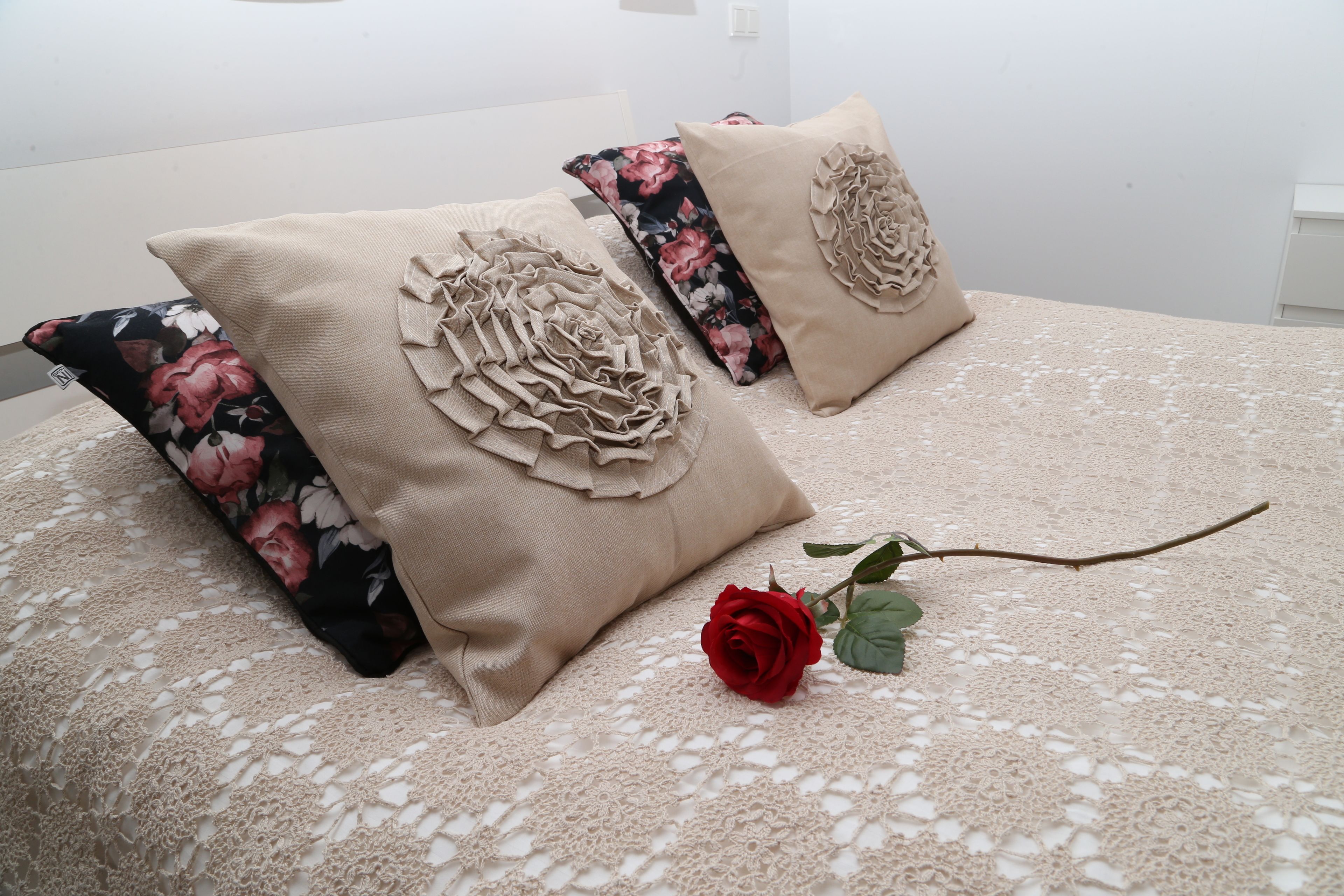 Una cama con un cojín estampado con motivos florales, un cojín color beige con un diseño decorativo y una rosa roja sobre la colcha.