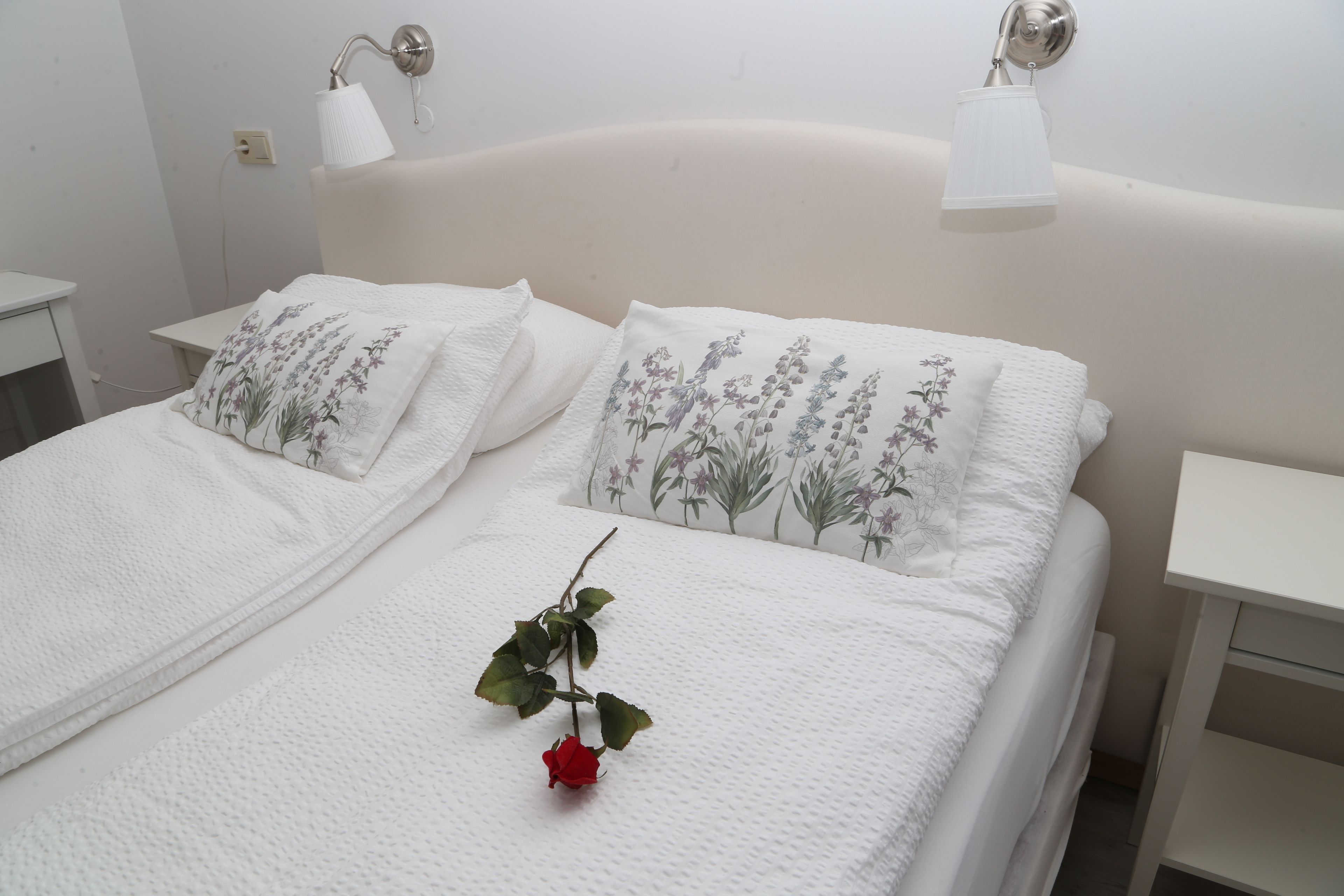 A neatly made bed with white bedding and floral pillows, a single rose on the bed, and bedside tables with lamps.