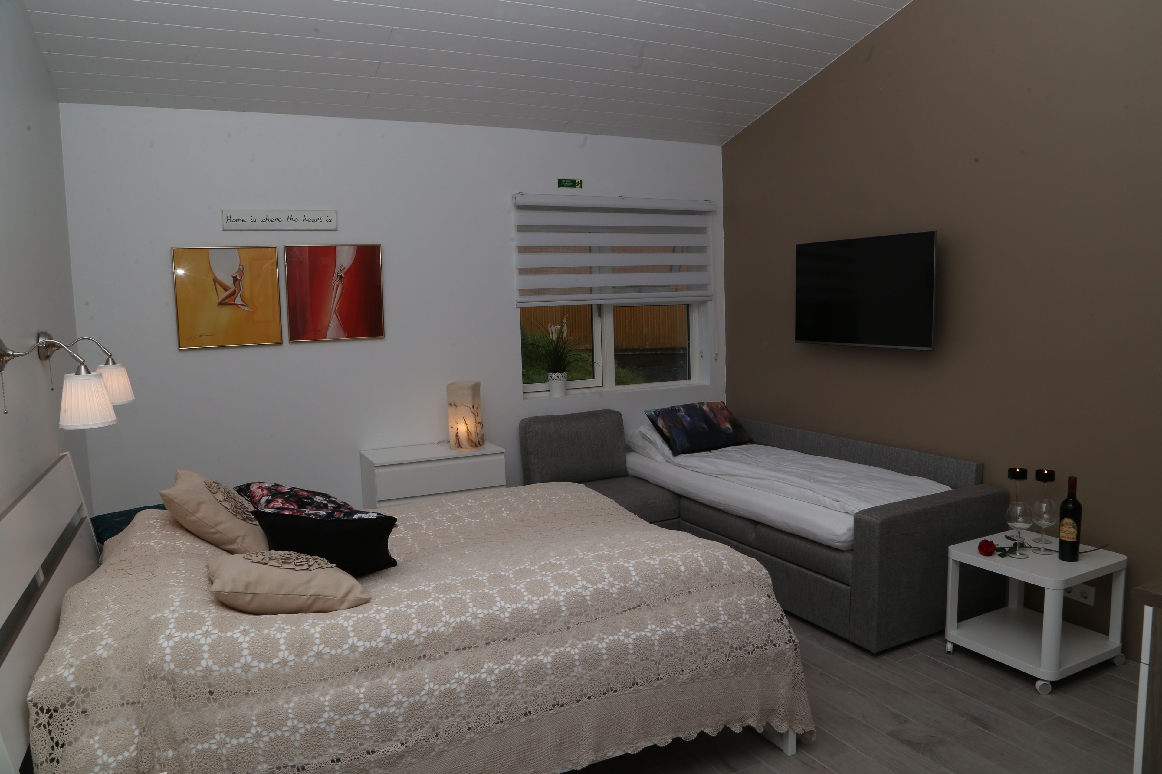 A bedroom with a large bed, a smaller bed, a TV, a bedside table with a lamp, and artwork on the wall.