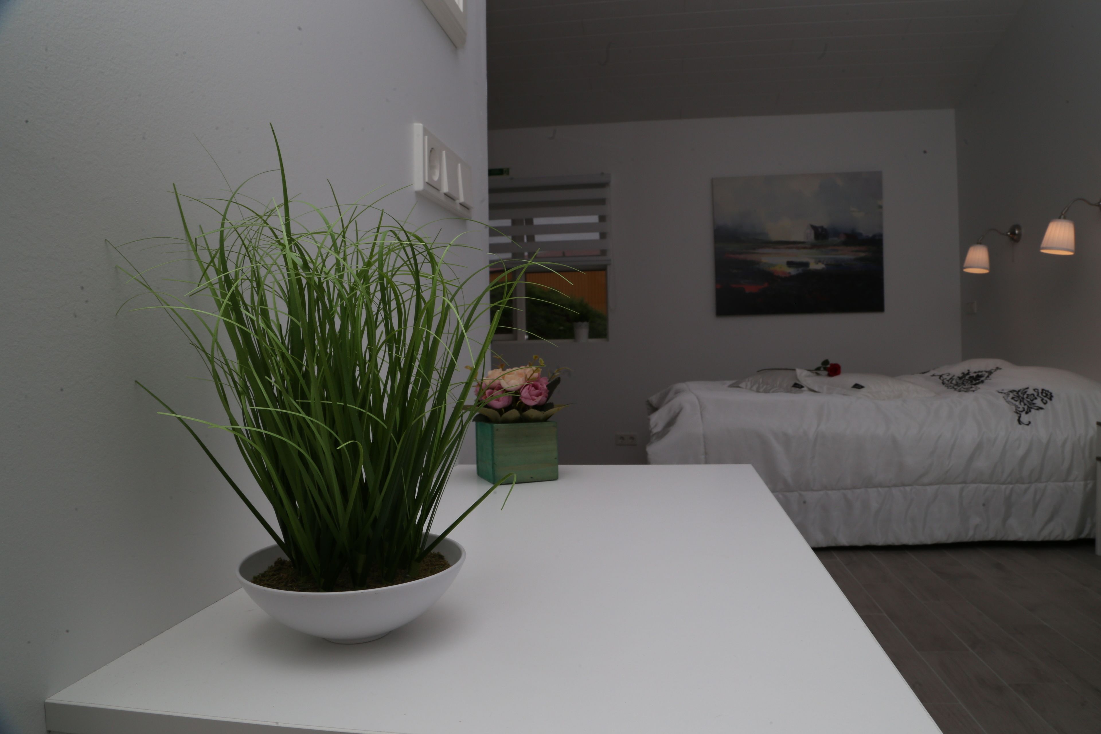 Un dormitorio moderno con una cama blanca, una pequeña planta en maceta y un cuadro en la pared.