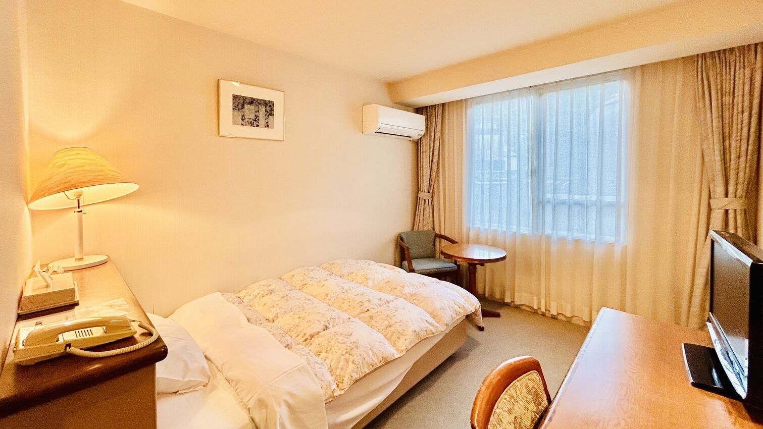 Ein Hotelzimmer mit einem Bett, einem Schreibtisch mit Computer, einem Stuhl, einer Lampe, einer Klimaanlage und einem fenster mit Vorhängen.