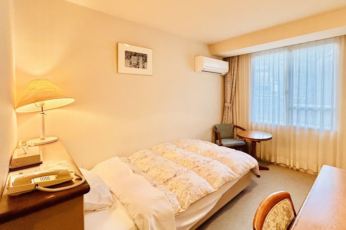 A hotel room with a bed, a desk with a computer, a chair, a lamp, an air conditioner, and a window with curtains.
