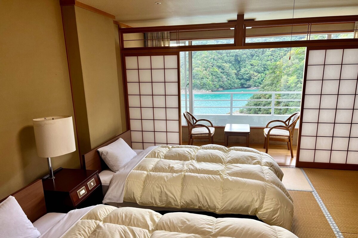 A hotel room with two beds, a nightstand, a lamp, a view of the sea, and a traditional Japanese sliding door.
