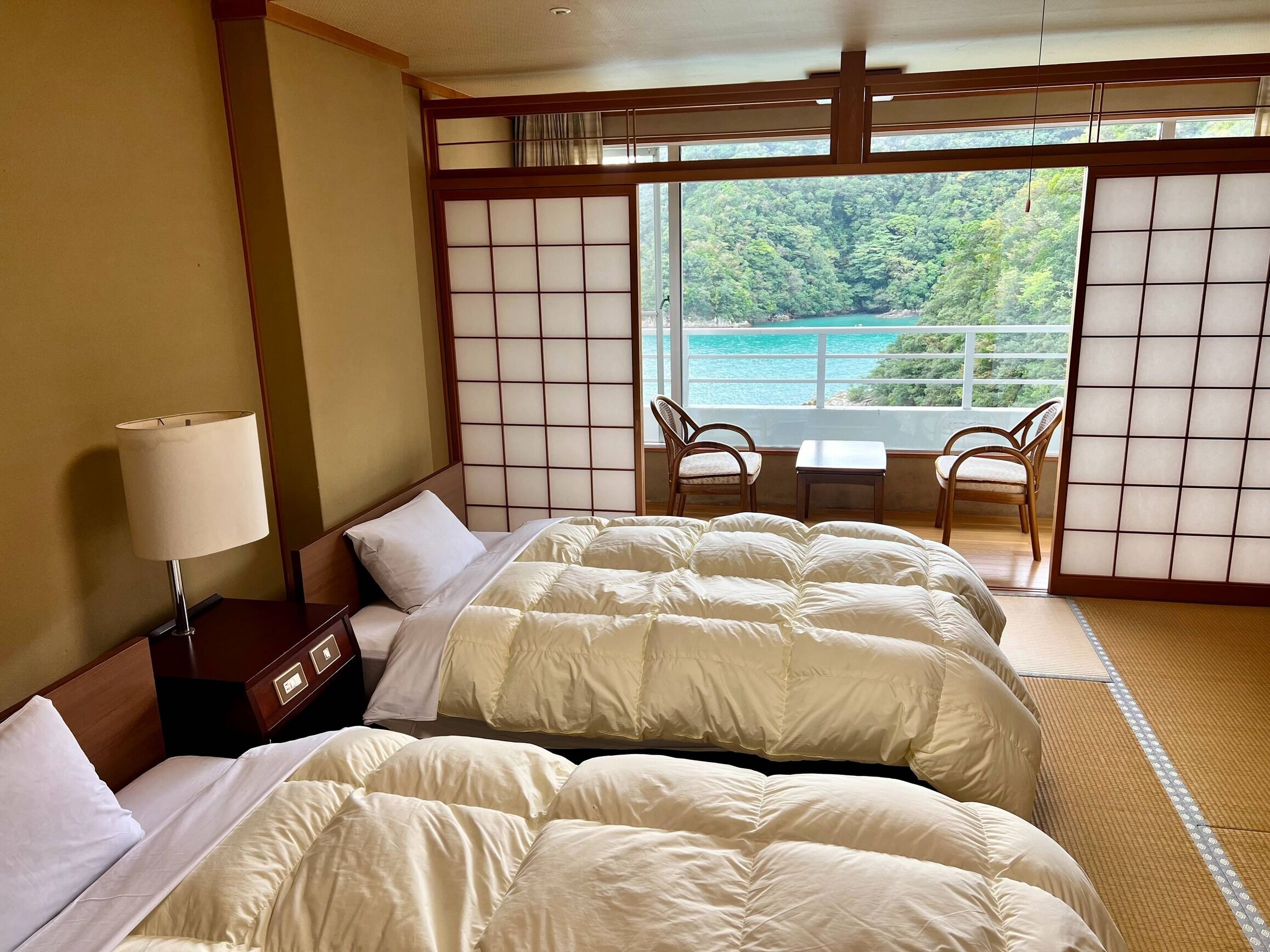 Ein Hotelzimmer mit zwei Betten, einem Nachttisch, einer Lampe, Blick auf das Meer und einer traditionellen japanischen Schiebetür.