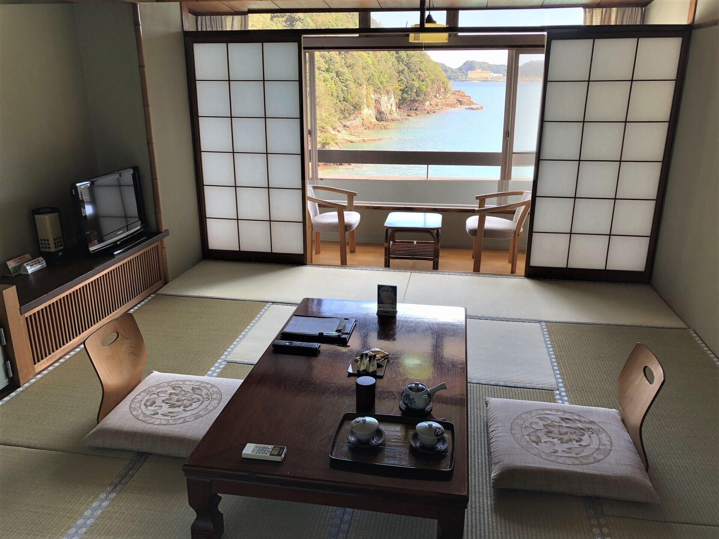A traditional Japanese room with tatami flooring, a low table with tea set, a television on a cabinet, and a view of a coastal landscape.