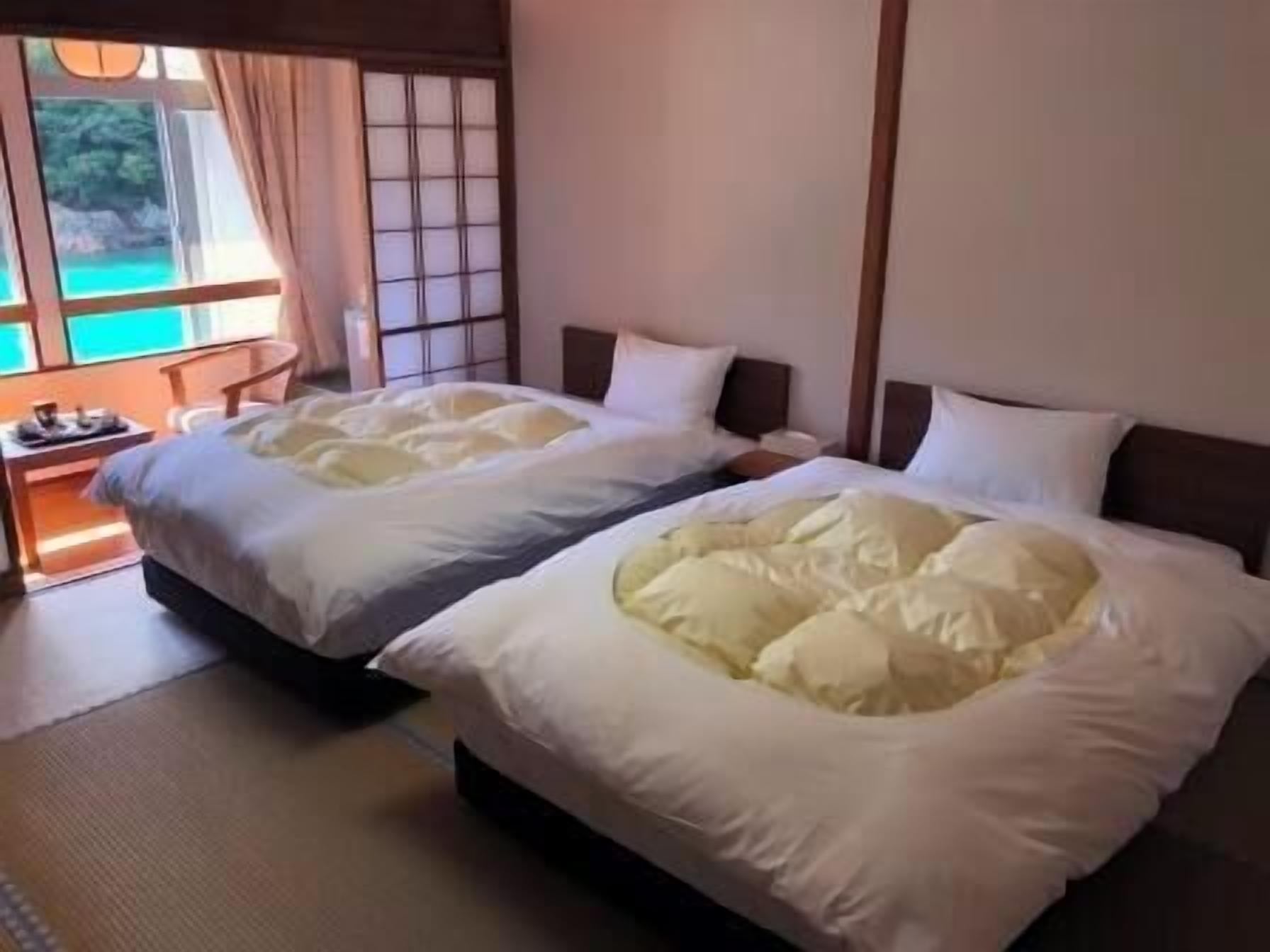 A hotel room with two beds, a wooden headboard, a small table with a lamp, and a view of a natural landscape through a window.