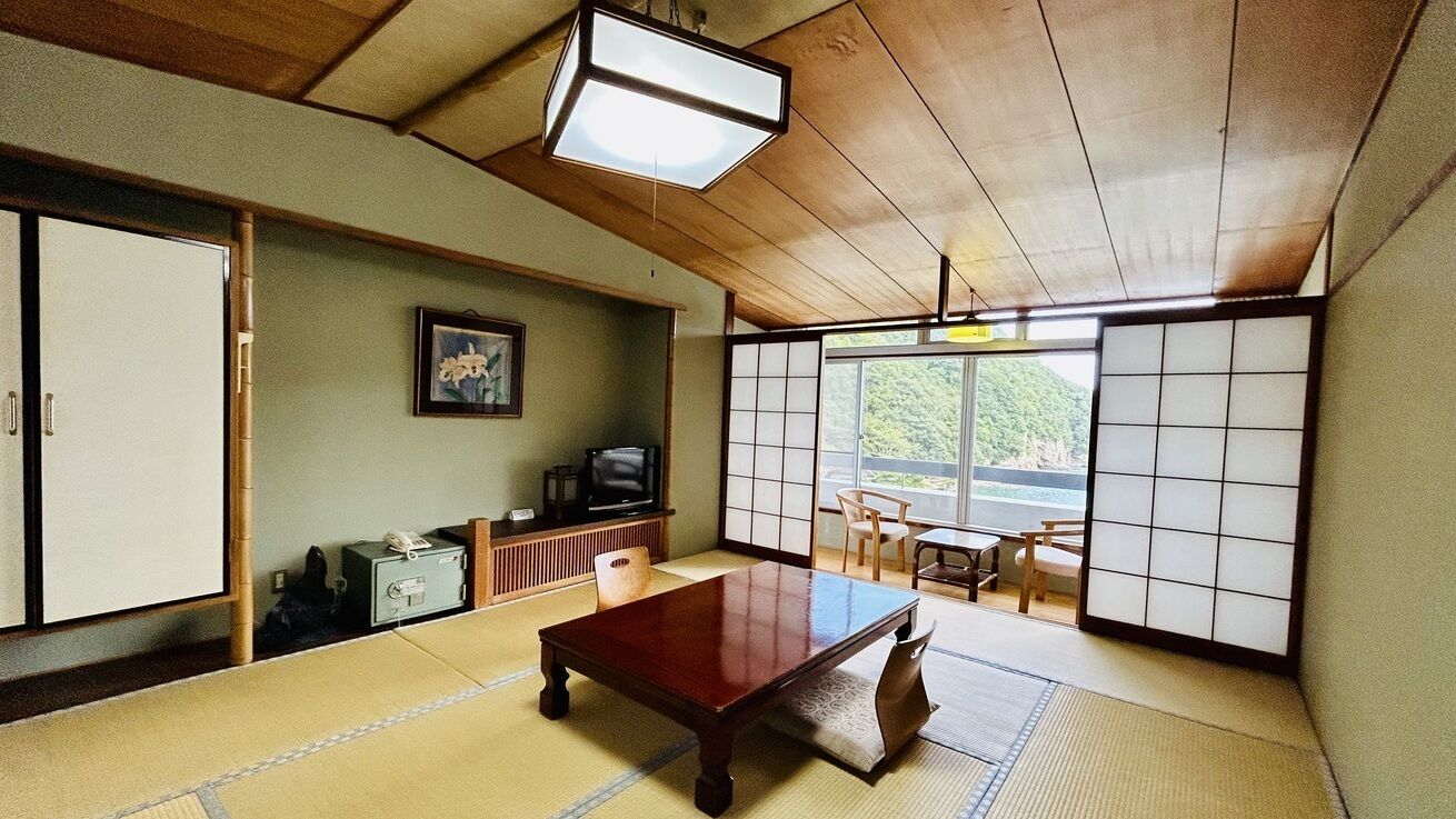 Ein traditioneller japanischer Raum mit Tatami-Boden, einem Holztisch, Schiebetüren, einem Fernseher und Blick ins Grüne.