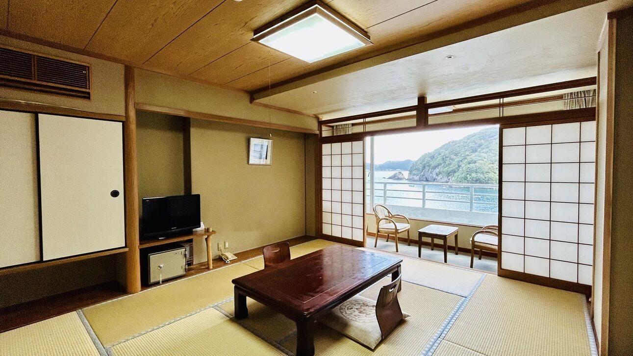 Ein traditioneller japanischer Raum mit Tatami-Böden, einem Holztisch, einem Fernseher und Schiebetüren, die auf einen Balkon mit Blick auf ein Gewässer und Hügel führen.