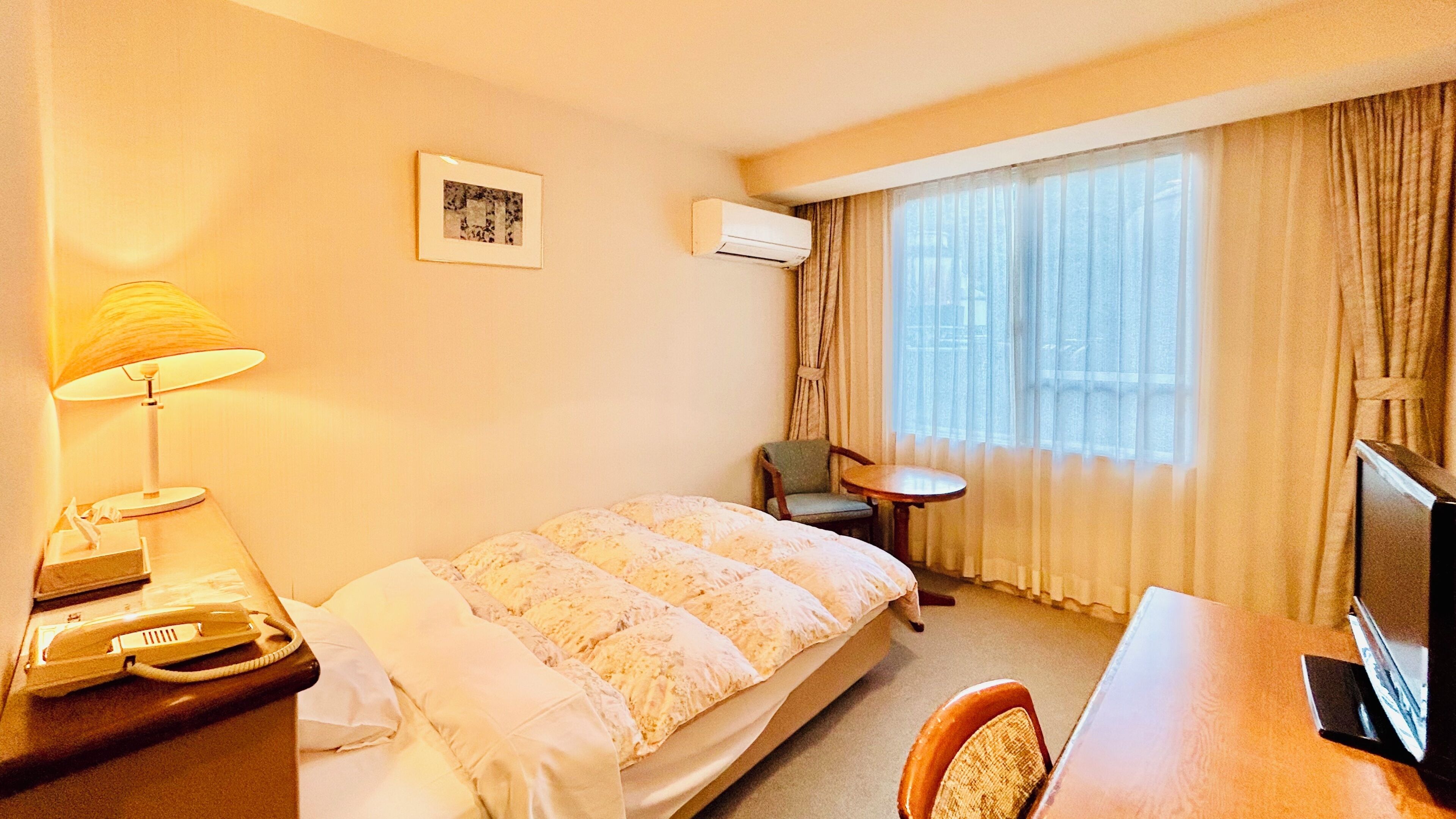 A hotel room with a bed, a desk, a chair, a lamp, an air conditioner, and a window with curtains.