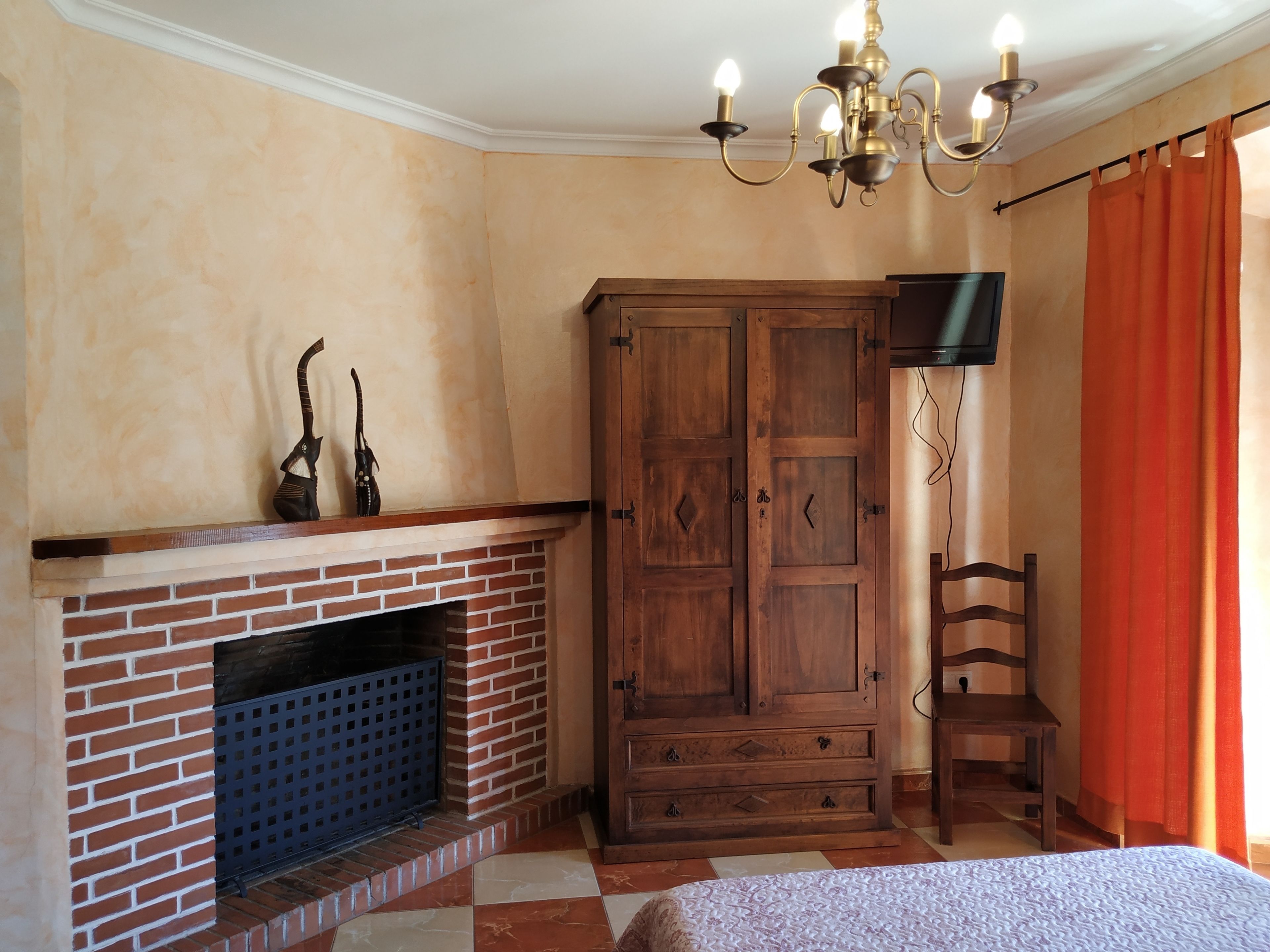 A bedroom with a brick fireplace, wooden wardrobe, a television mounted on the wall, and a bed with a patterned bedspread.