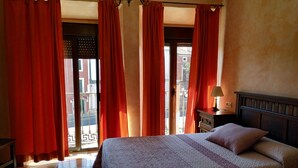 Habitación de hotel con cama, mesita de noche, lámpara y vista de la calle a través de amplias ventanas con cortinas rojas.
