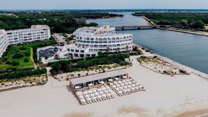 Vue aérienne d’un hôtel situé sur la côte, composé de plusieurs bâtiments, avec une plage équipée de transats et une rivière en arrière-plan.