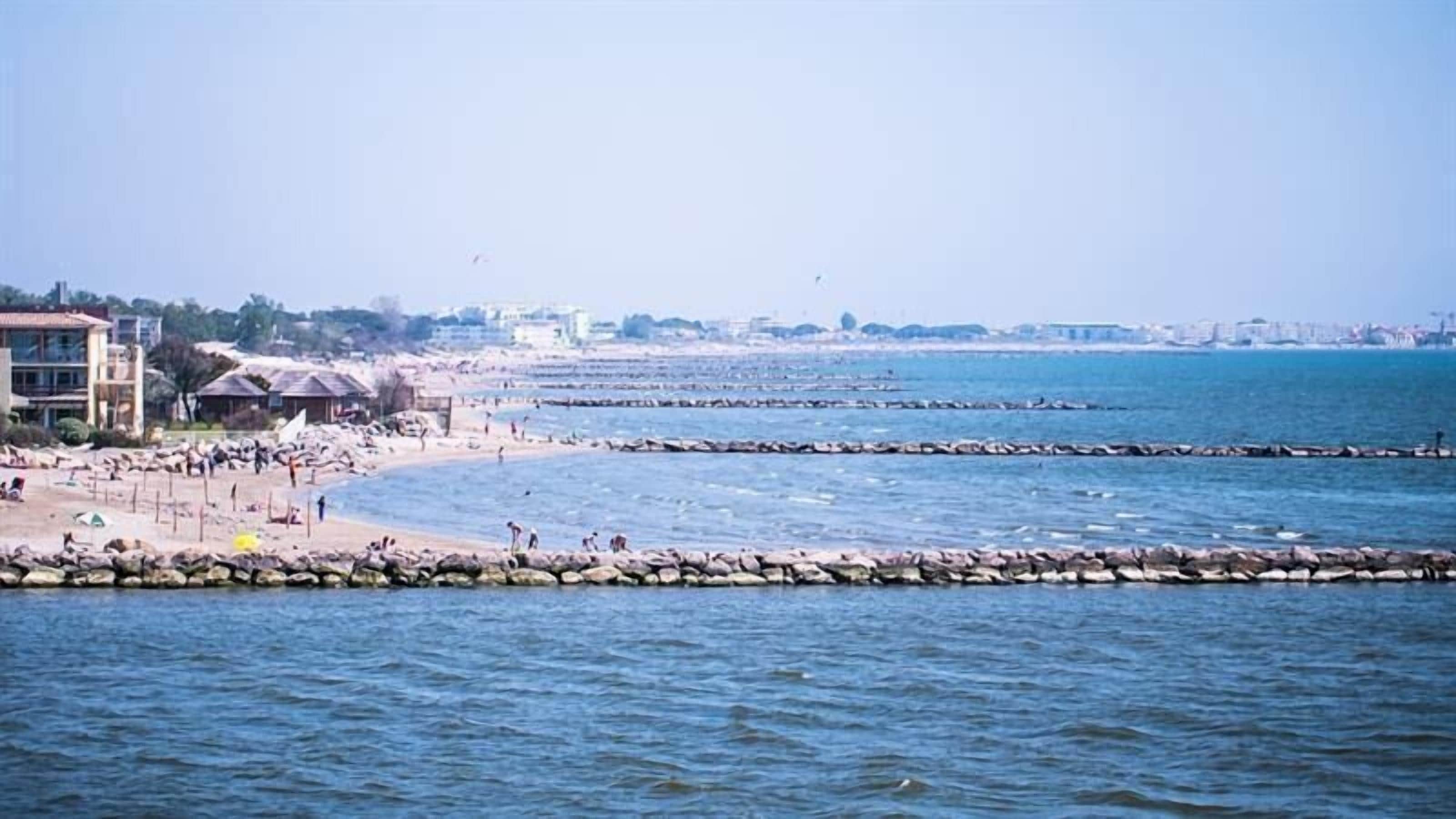 Une plage côtière avec des brise-lames rocheux, une étendue de sable et des bâtiments en arrière-plan.
