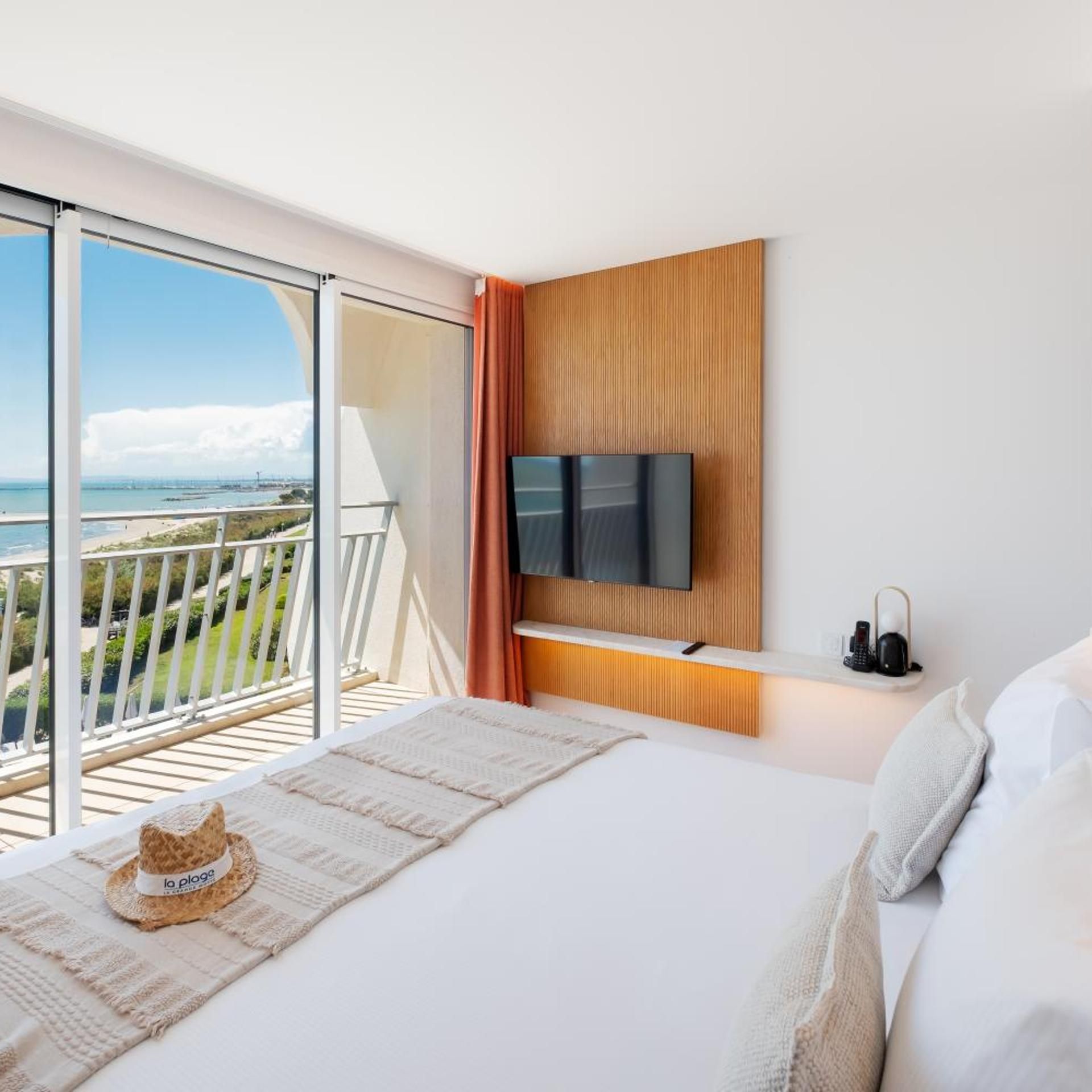 Une chambre d’hôtel avec un grand lit, un téléviseur fixé sur un panneau en bois, un balcon avec vue sur la mer et une petite table sur laquelle est posé un chapeau.