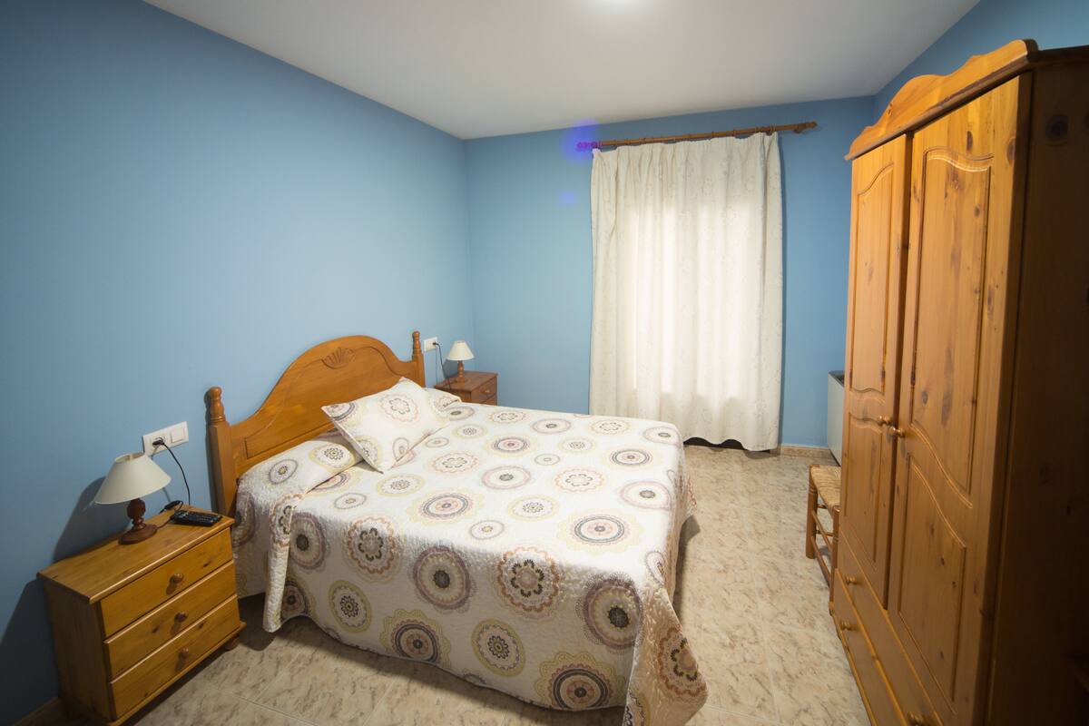 A bedroom with a wooden bed, a nightstand with a lamp, a wardrobe, and a window with curtains.