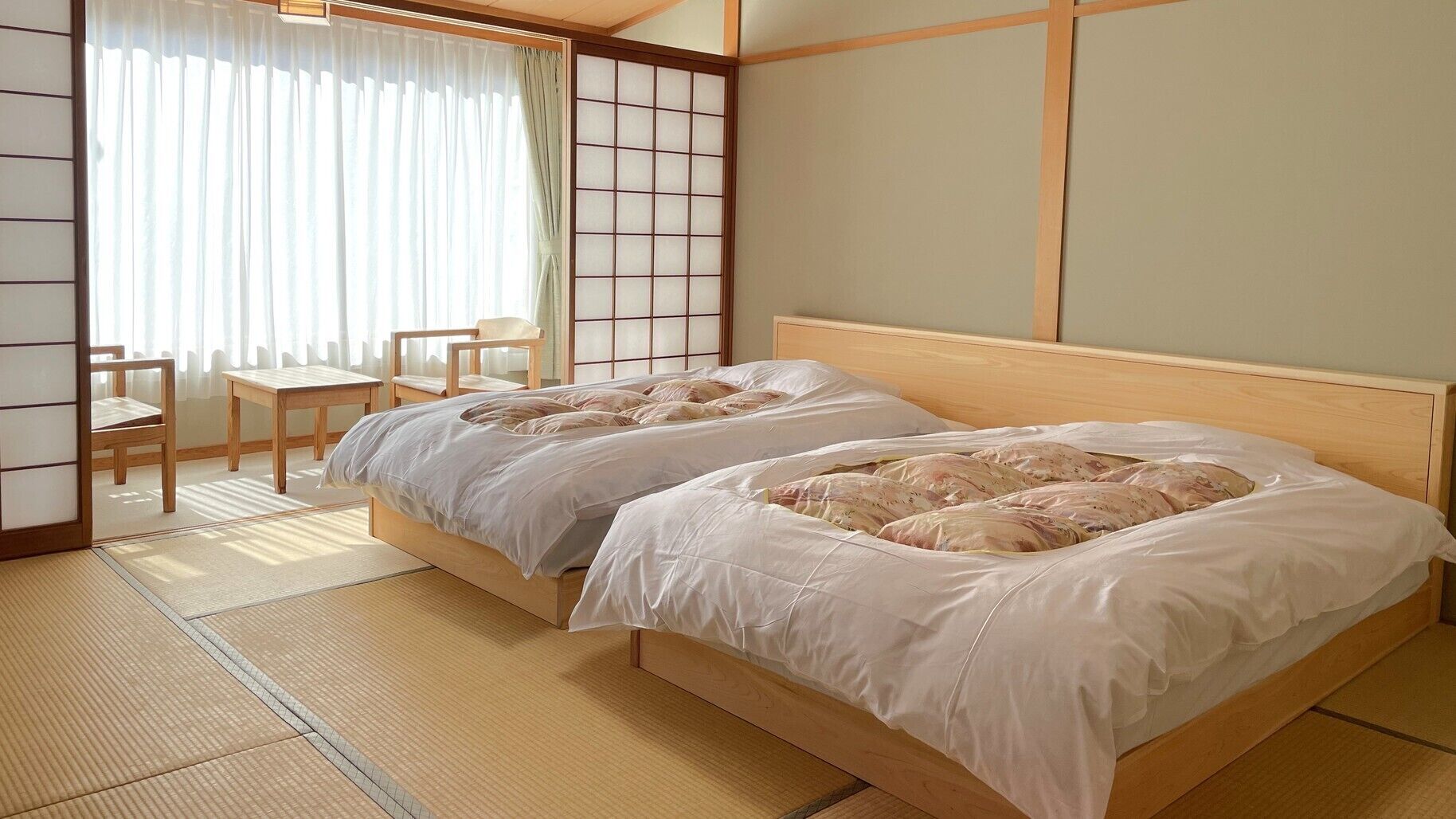 Una habitación tradicional japonesa con suelo de tatami, una cama baja de madera con ropa blanca, un shoji y una pequeña mesa con sillas.