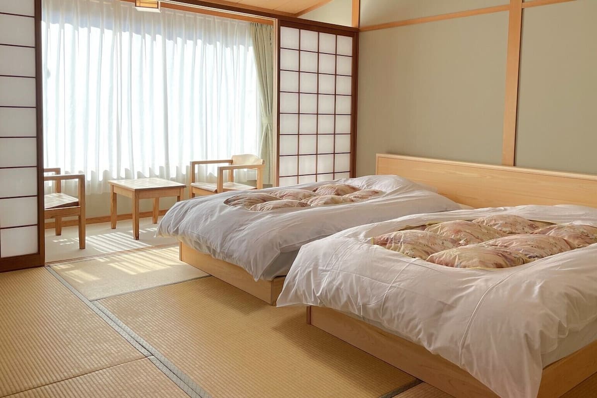 A traditional Japanese-style room with tatami flooring, a low wooden bed with white bedding, a shoji screen, and a small table with chairs.