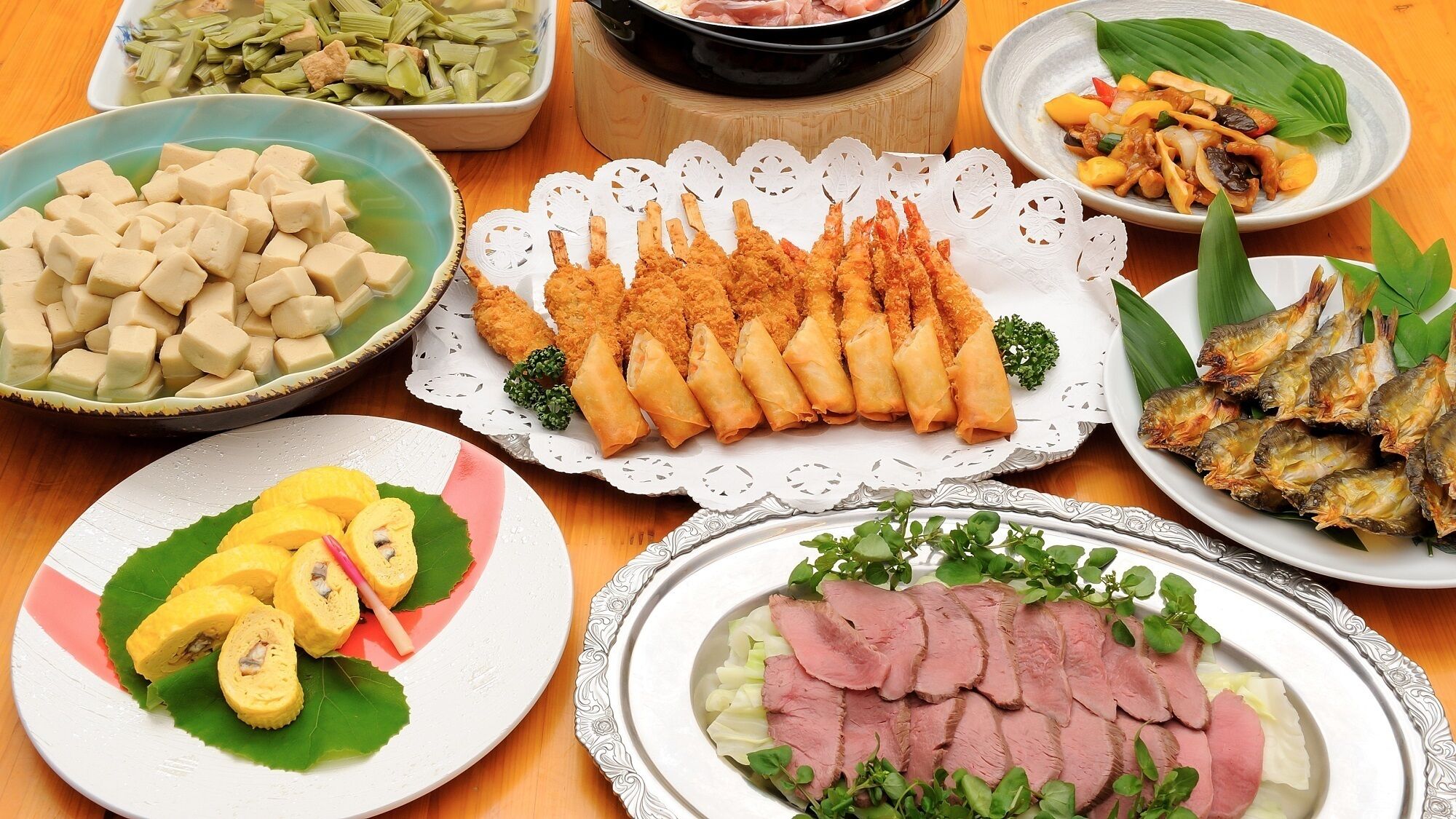 Una variedad de platos que incluyen tofu, rollitos primavera y pescado, dispuestos sobre una mesa de madera.