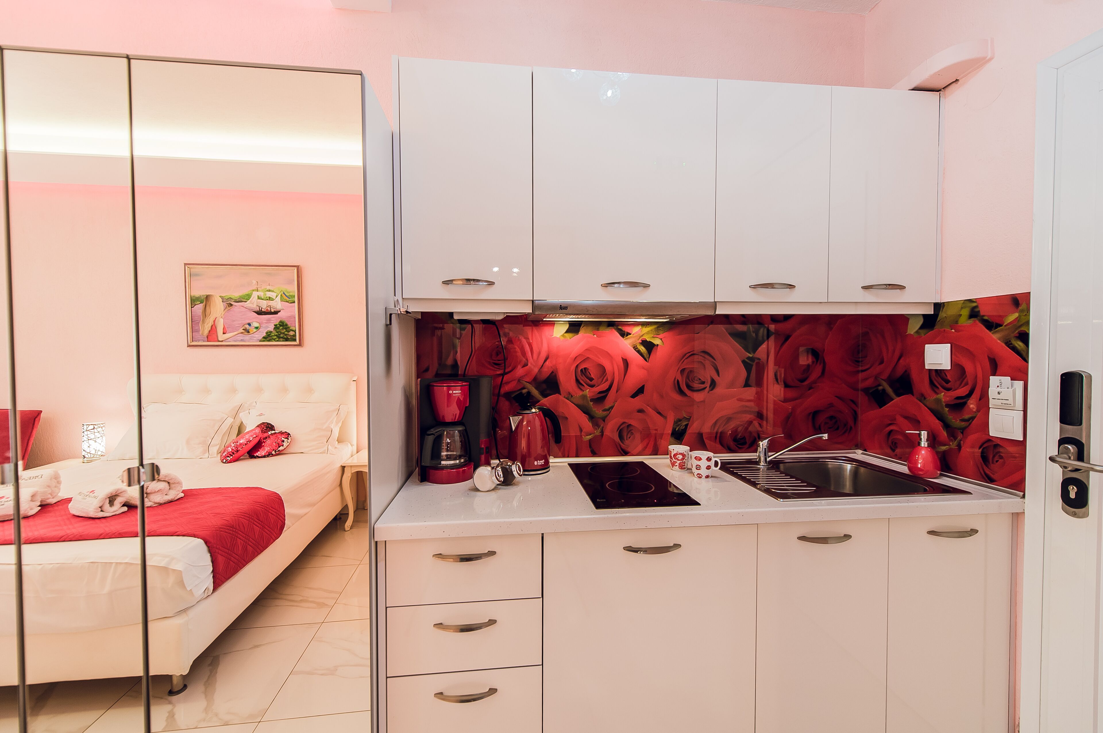 Un dormitorio con cama, una mesita de noche y un mueble auxiliar. La habitación tiene una pared rosa y un cuadro. La imagen también muestra una cocina con armarios blancos, un fregadero y una estufa.