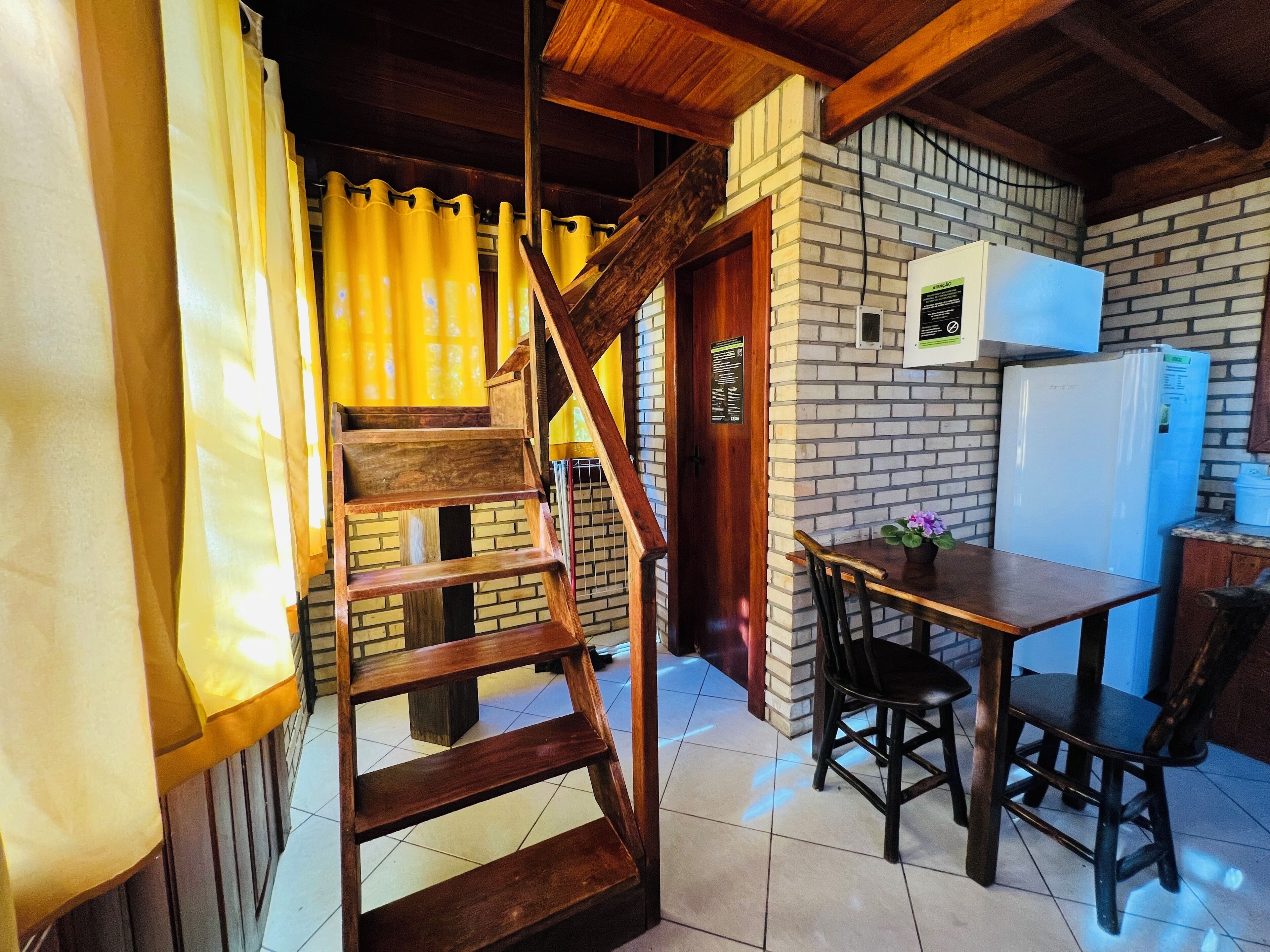 Un área de cocina con una escalera de madera, una mesa de comedor con sillas, un refrigerador y una pared de ladrillo.