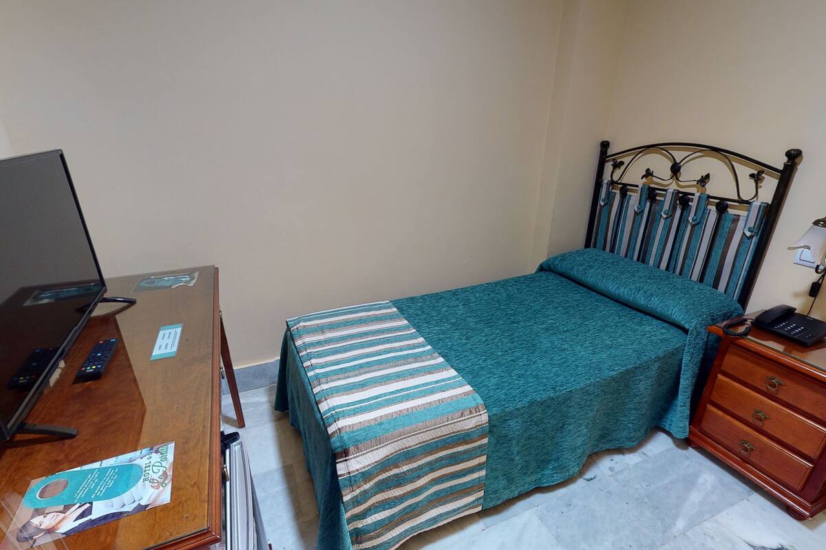 A bedroom with a bed, a desk with a laptop, a telephone, and a bedside table.