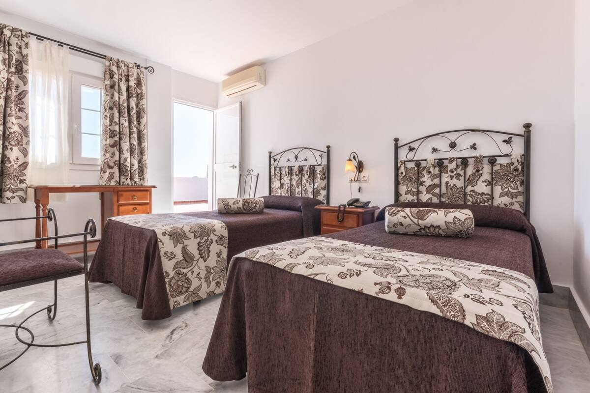 A bedroom with two beds, a desk, a bench, and a window with curtains.