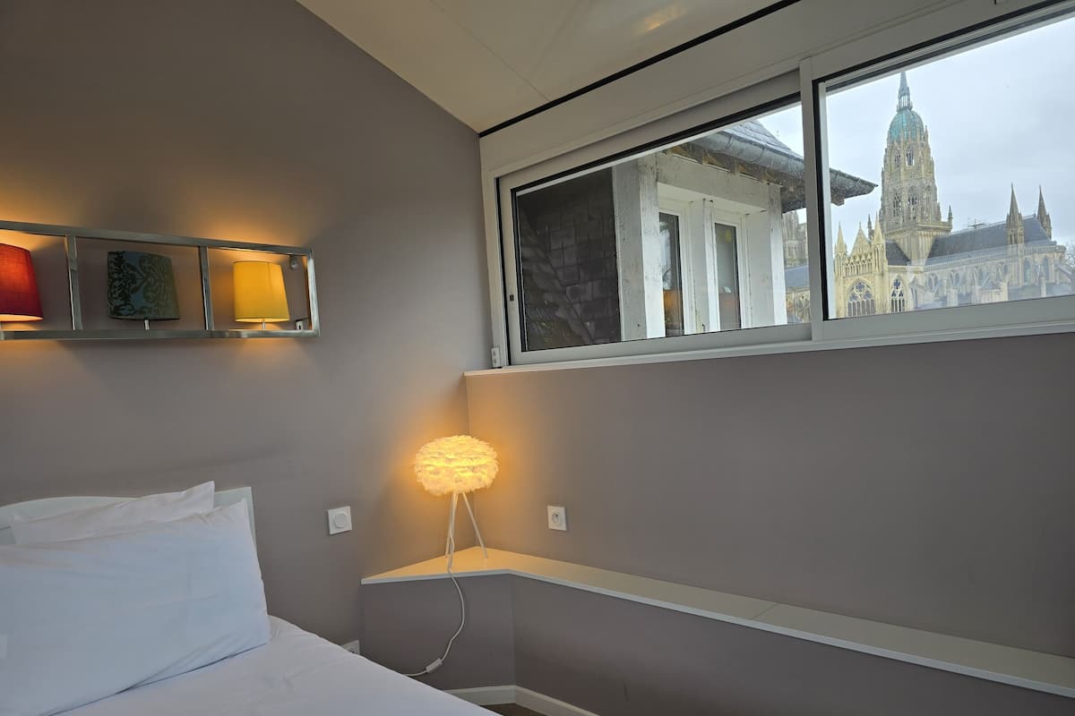 A hotel room with a bed, bedside lamps, a window with a view of a cathedral, and a shelf with a lamp.