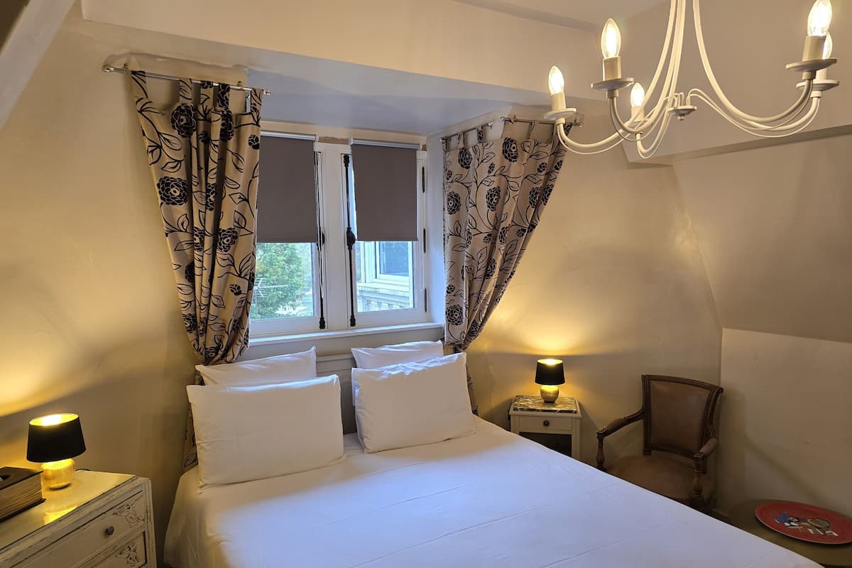 A bedroom with a bed, a nightstand with a lamp, a chair, and a window with curtains.