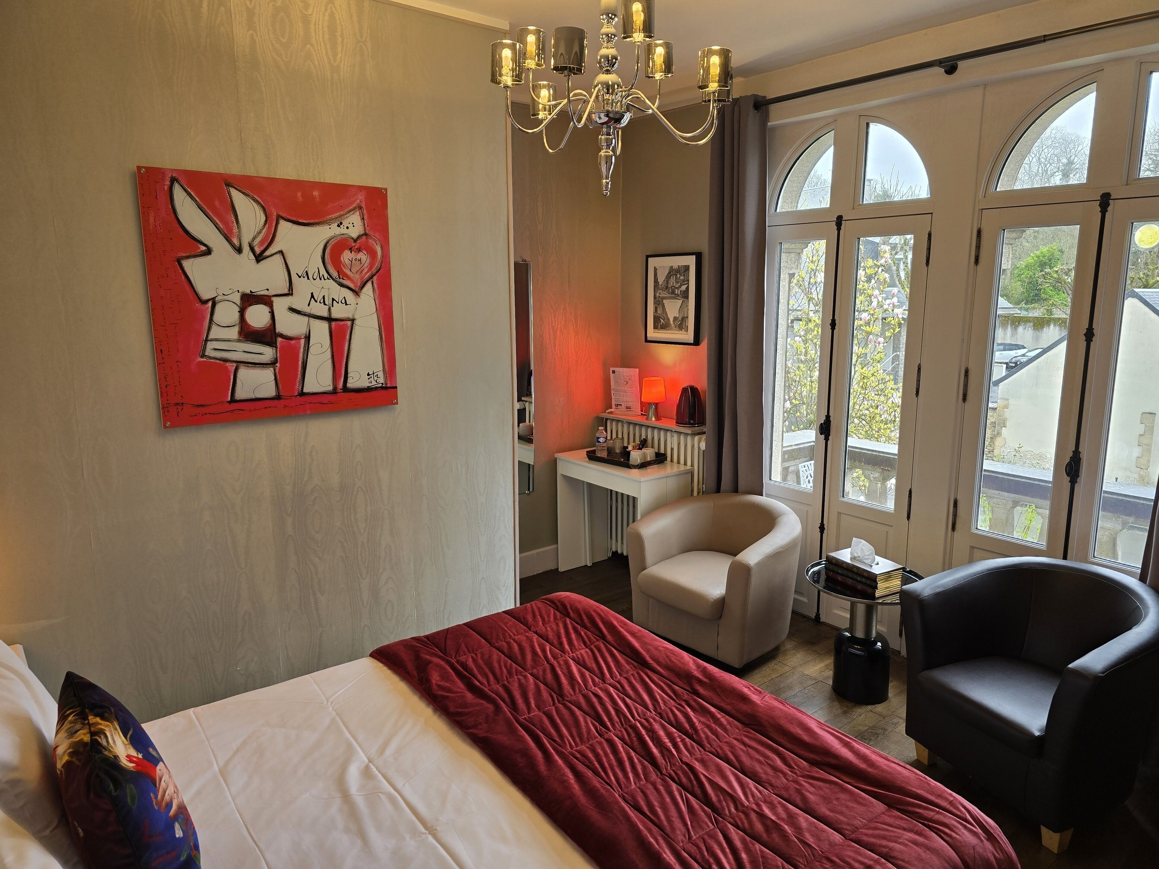 Habitación de hotel con cama, escritorio con lámpara, silla, manta roja, candelabro y ventana grande con vistas al exterior.