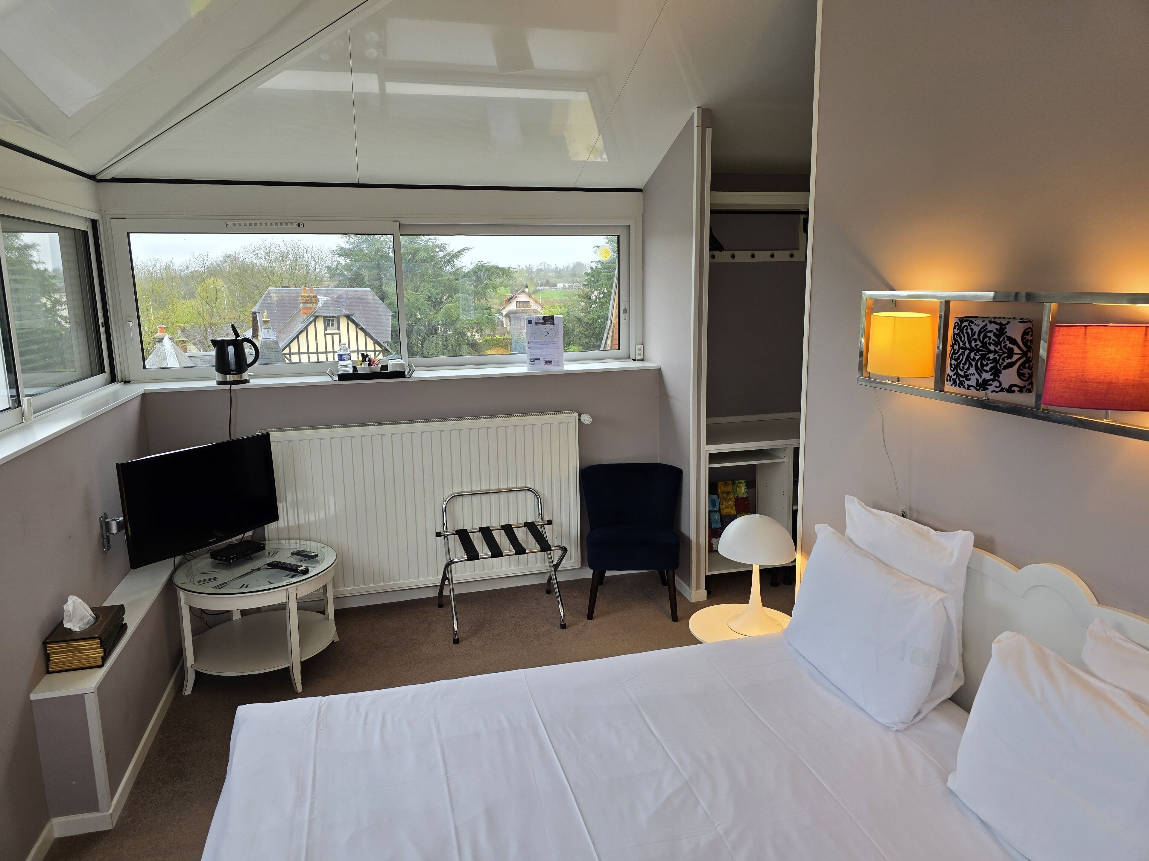 A hotel room with a bed, a desk with a TV, a chair, a radiator, a window with a view of houses, and a shelf with lamps.