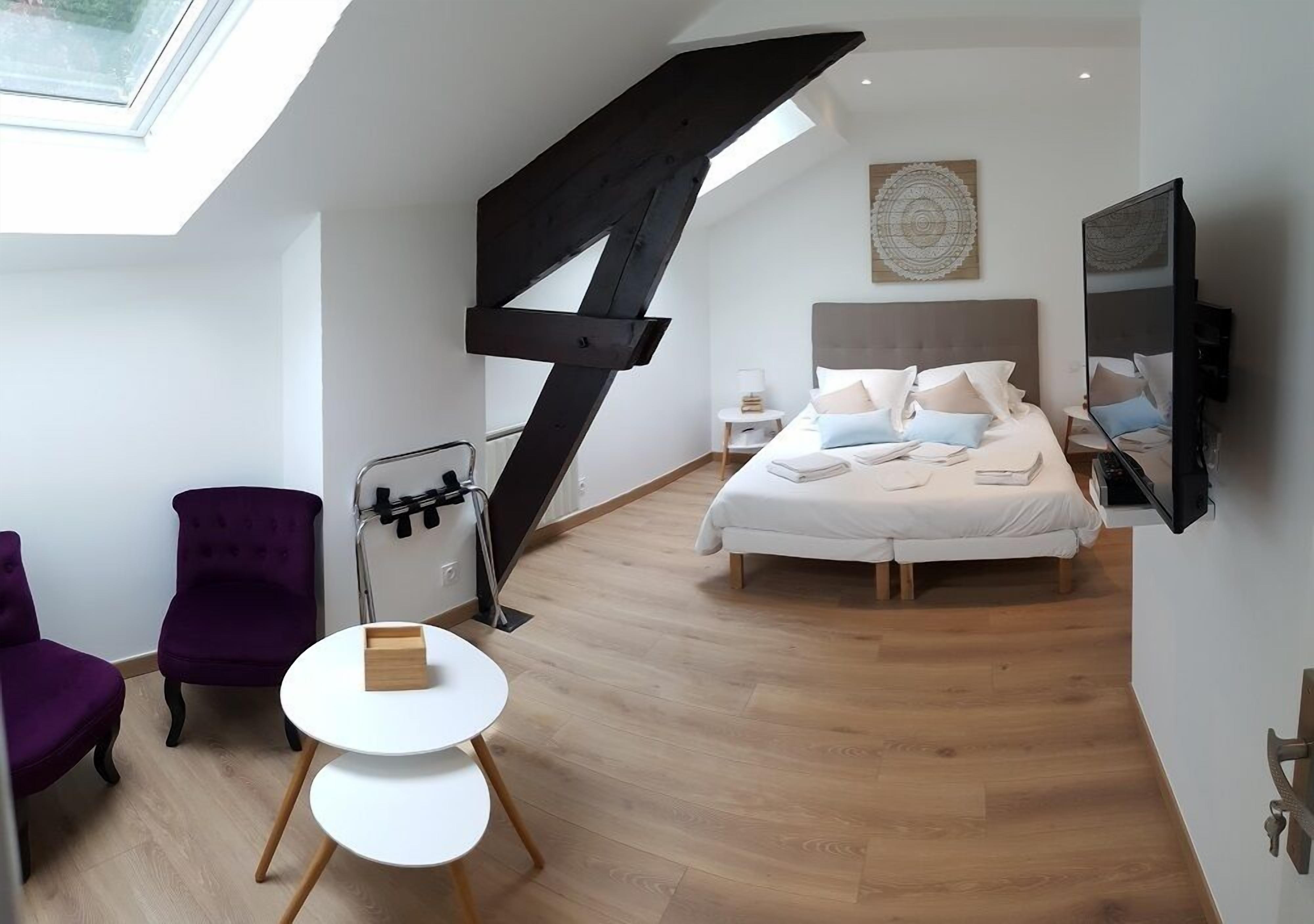 A modern bedroom with a skylight, a bed with pillows, a small round table, and purple chairs.