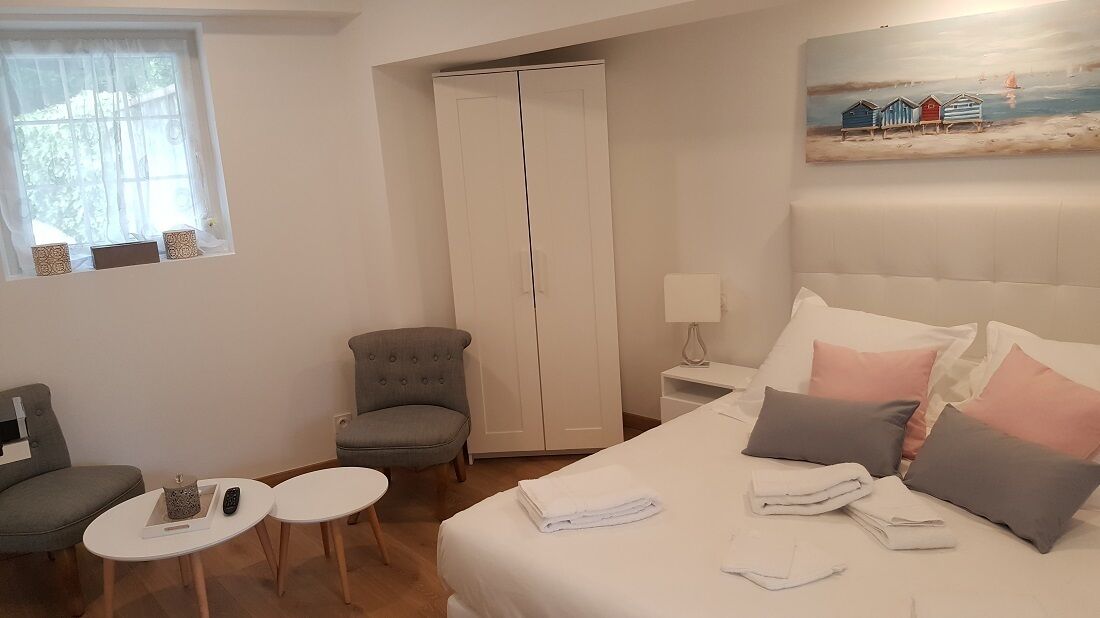 A hotel room with a bed, a grey armchair, a small table, a wardrobe, and a painting on the wall.