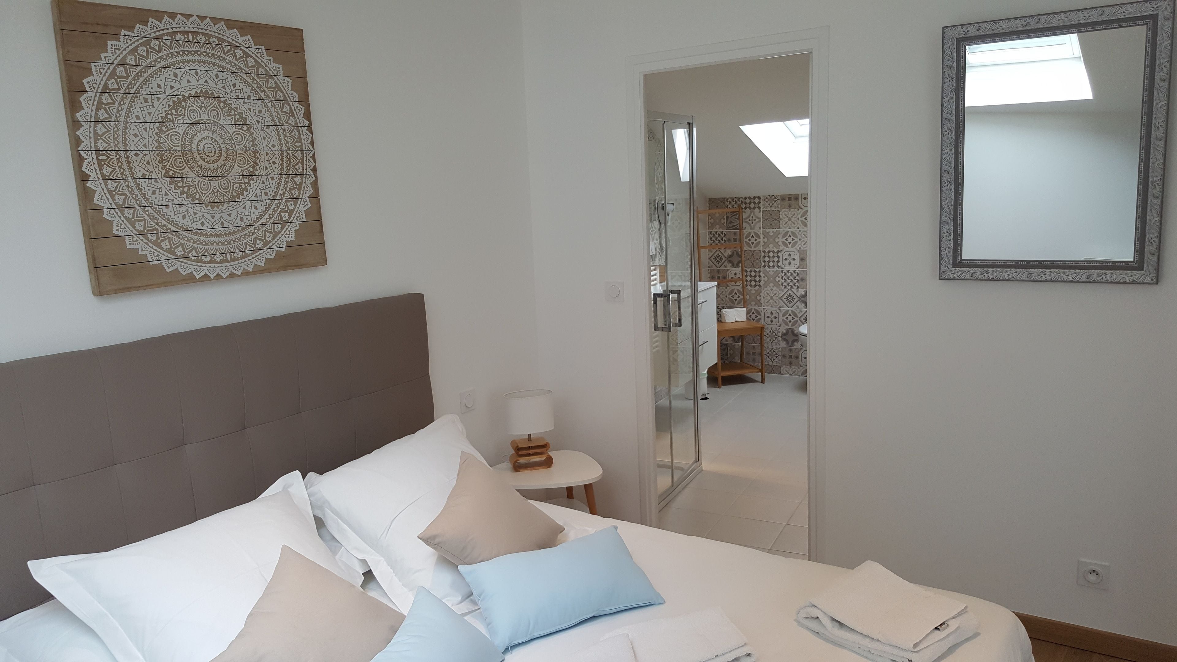 A bedroom with a bed, pillows, a bedside table, a mirror, and a wall with a decorative piece.