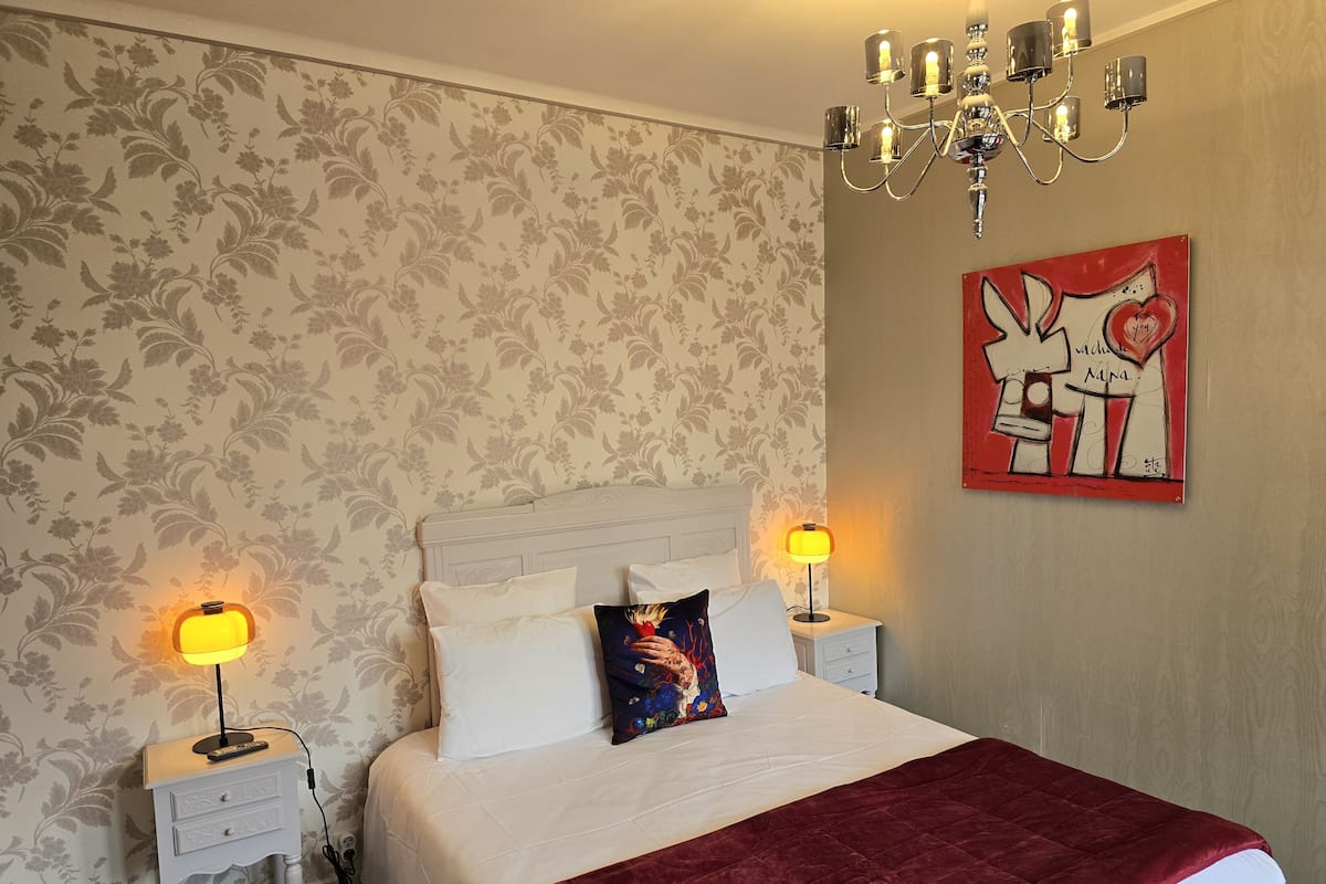 A bedroom with a bed, two bedside tables, a chandelier, a wall with floral wallpaper, and a piece of art.