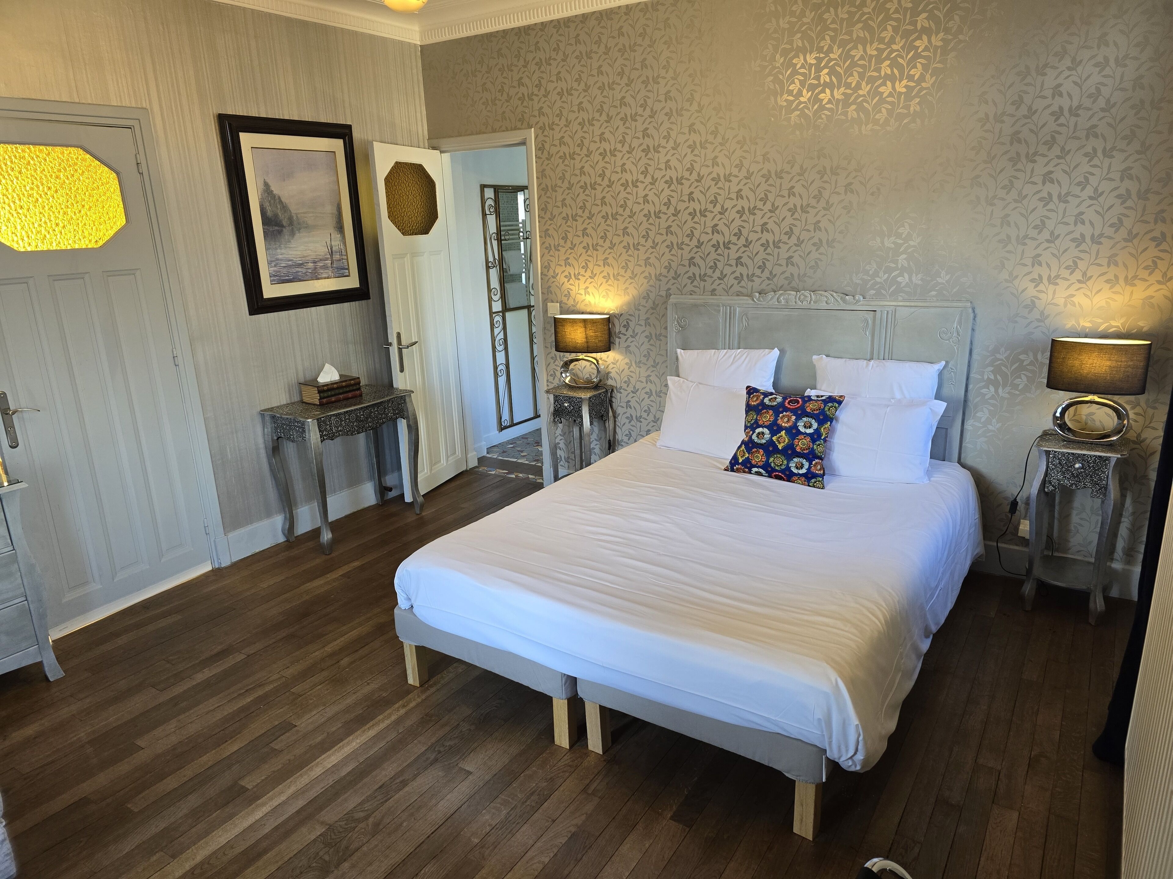 A neatly arranged bedroom with a large bed, two bedside tables with lamps, a framed picture on the wall, and a door with a textured glass panel.