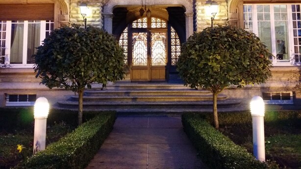 Un edificio imponente con una fachada simétrica, una entrada central con una puerta decorativa y dos árboles bien cuidados a cada lado de la entrada.