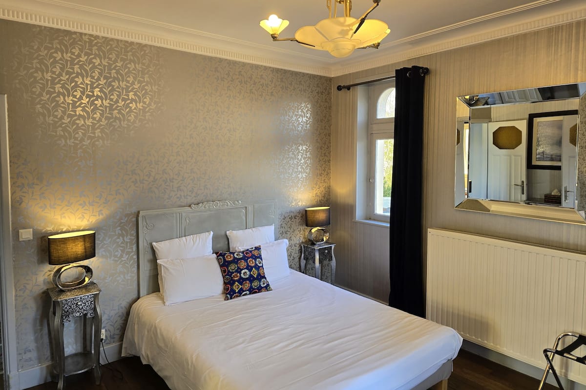 A neatly arranged bedroom with a large bed, two bedside tables with lamps, a mirror, and a window with curtains.