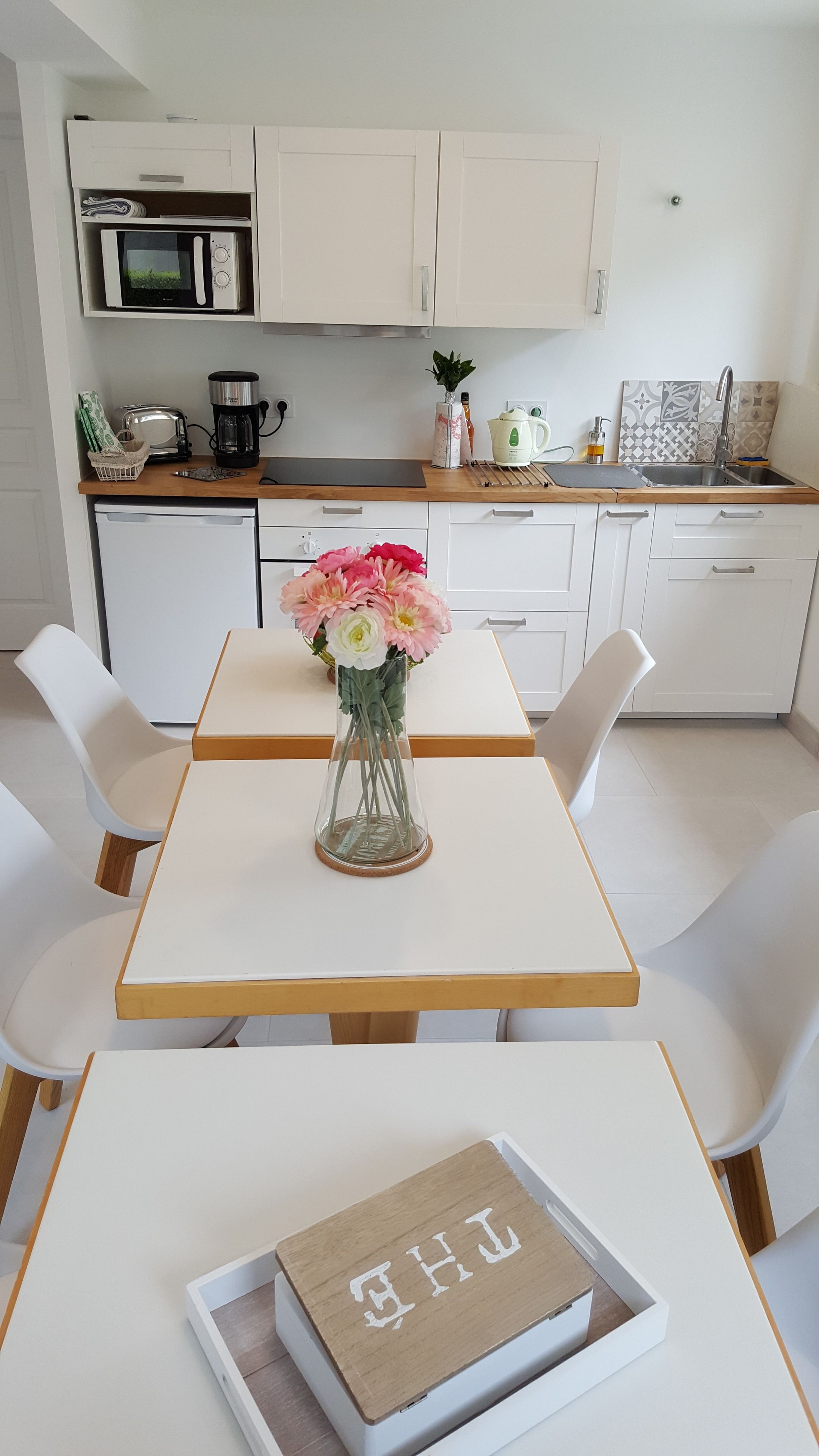 Een moderne keuken met witte kastjes, een magnetron, koffiezetapparaat en een spoelbak. Er staat een tafel met stoelen en een vaas met bloemen.