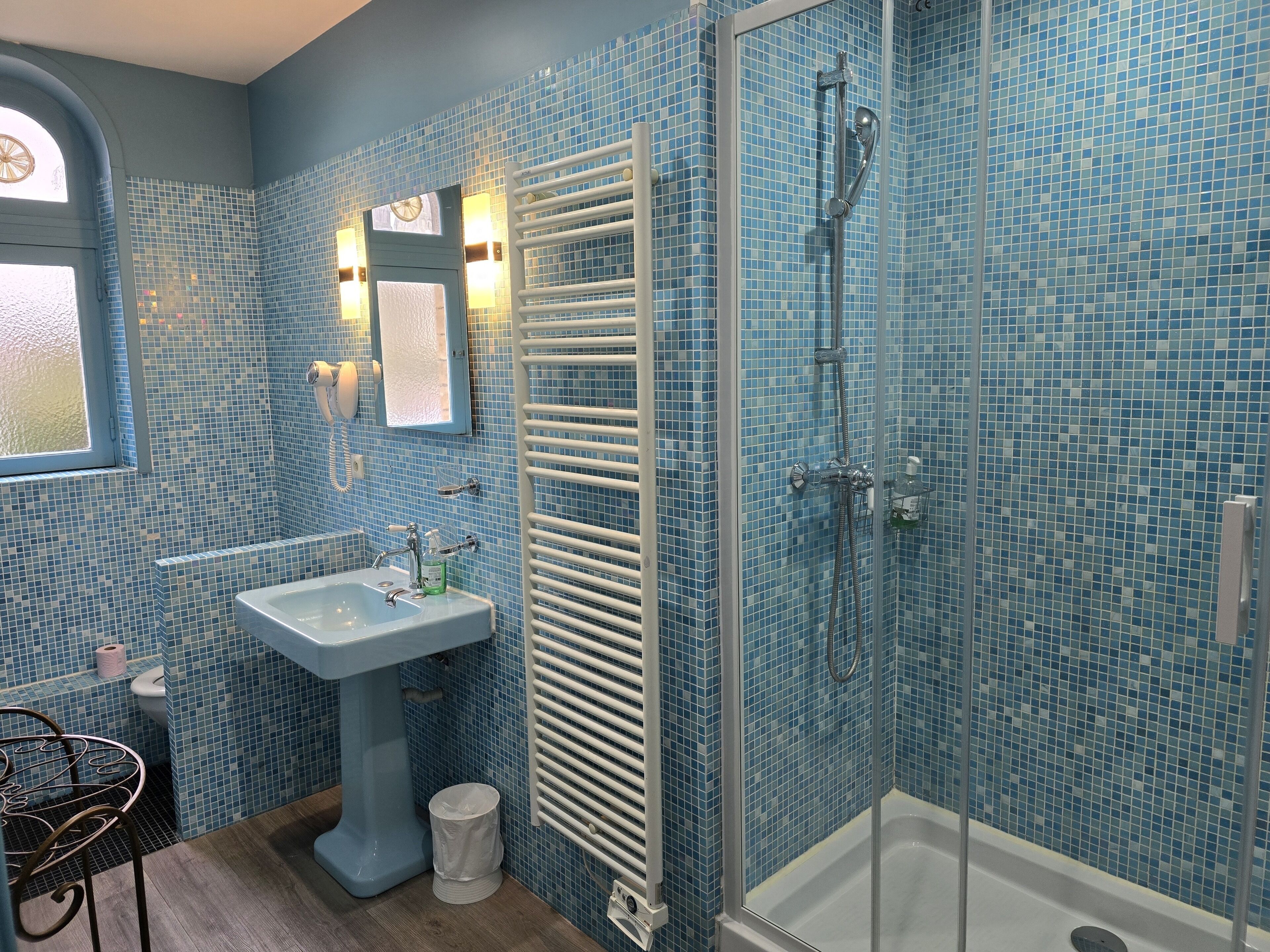 A bathroom with a blue mosaic tile wall, a pedestal sink, a glass shower enclosure, and a window with a view.